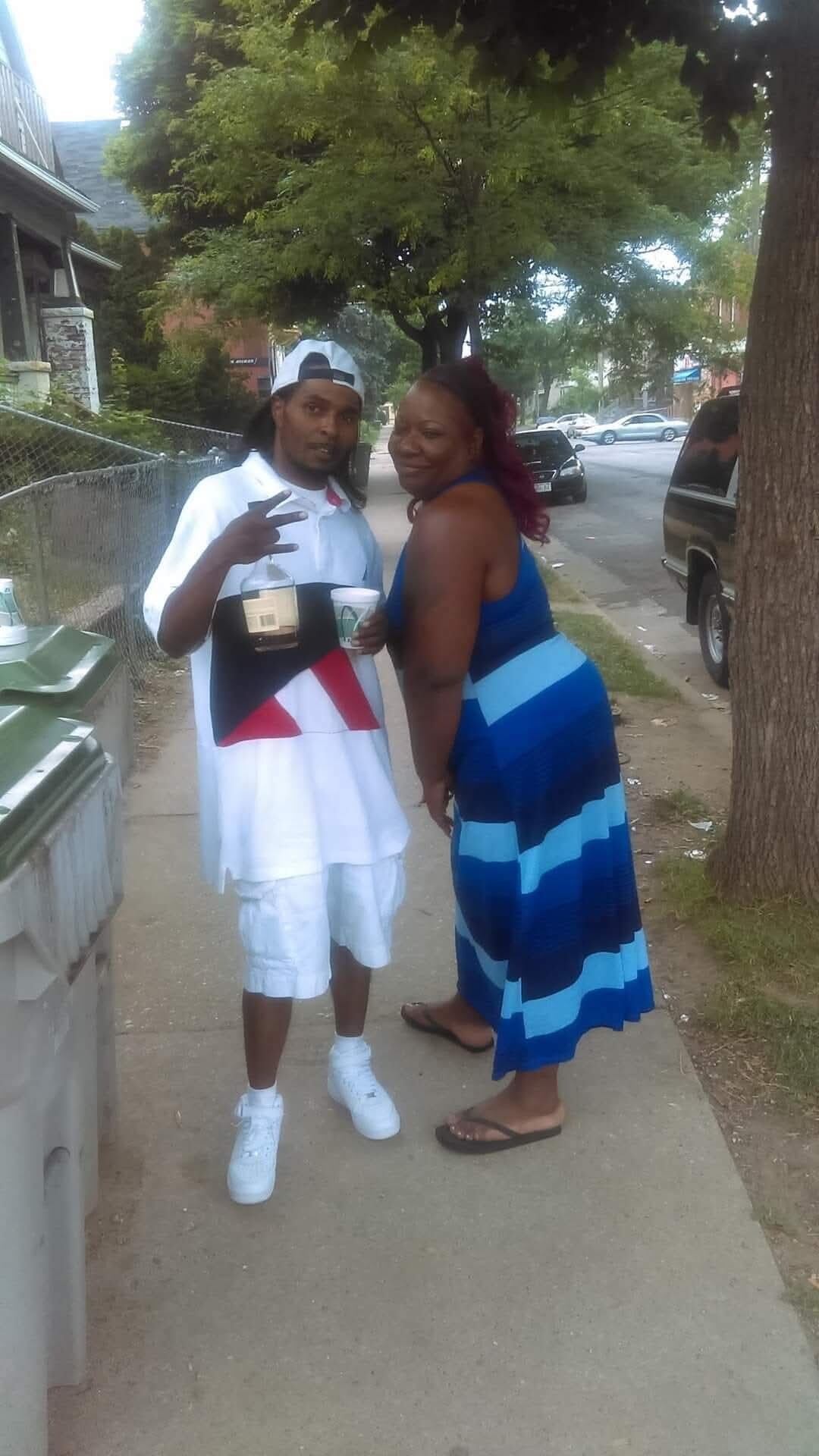 Two friends posing on a sidewalk; man in white streetwear and cap holding a bottle and cup, making a peace sign, with a woman in a blue striped dress smiling next to him.