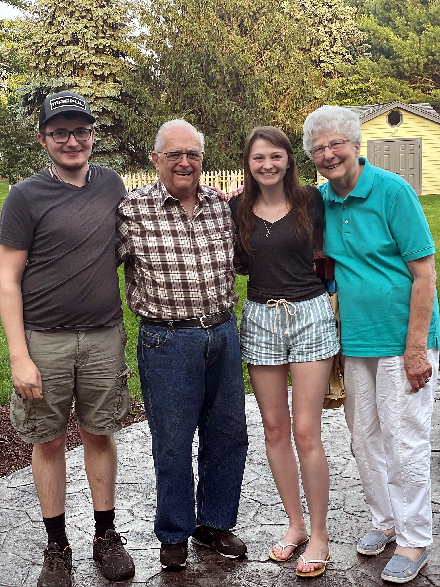 Group of four smiling people standing outdoors in a yard with trees and a fence behind them, posing for a photo.