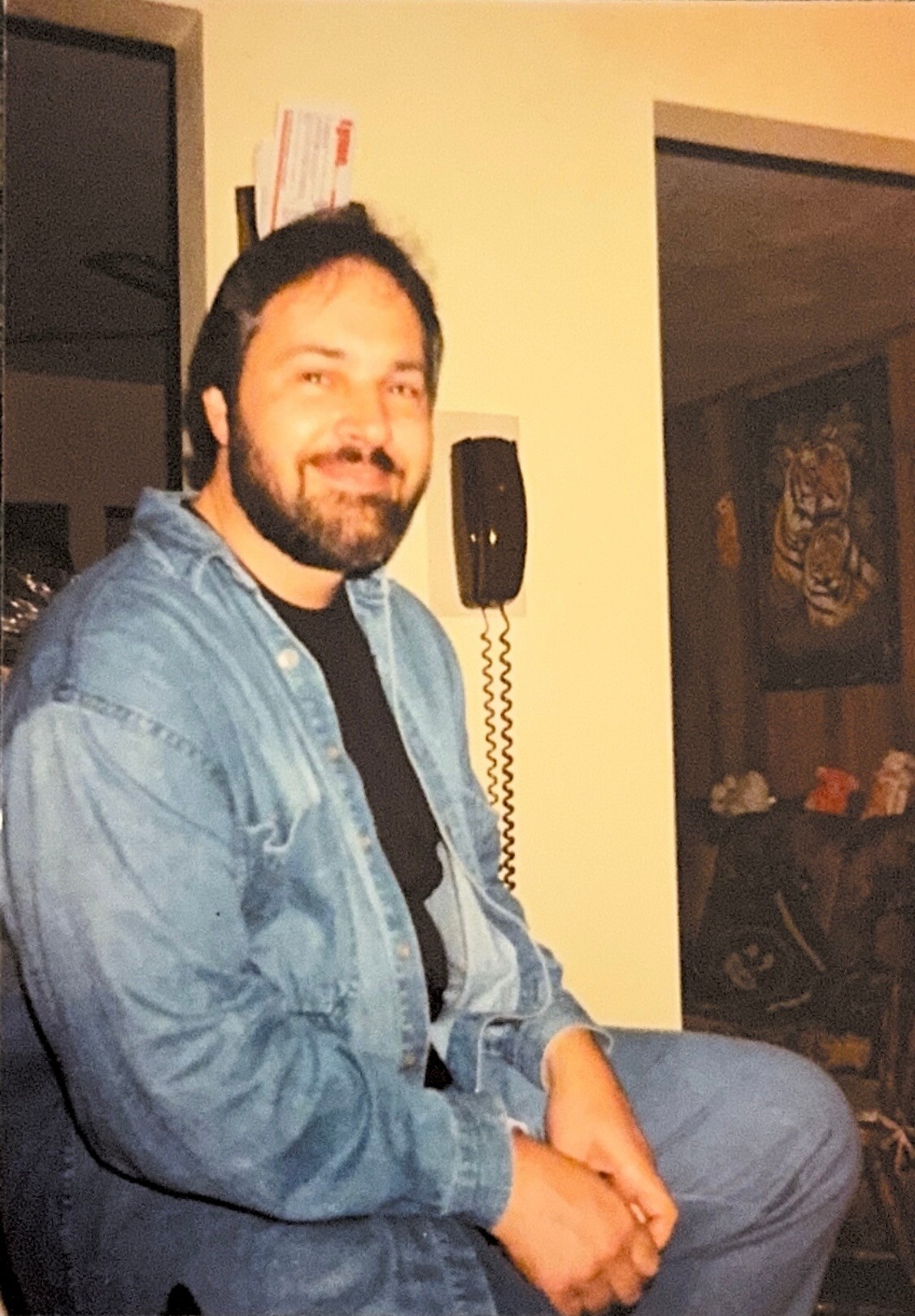 Smiling man with dark hair and beard, wearing a denim shirt over a black tee, seated indoors near a wall-mounted phone.