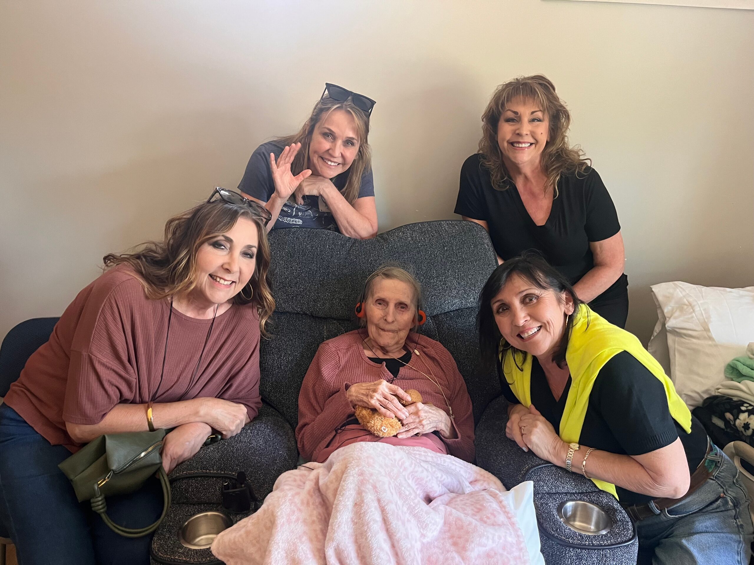 Six women of various ages sitting on a couch, smiling at the camera; elderly woman wrapped in a pink blanket.
