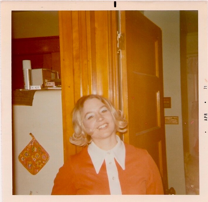 Young woman with blonde hair in a red blazer and white collar smiles at the camera indoors, standing by a wooden door.