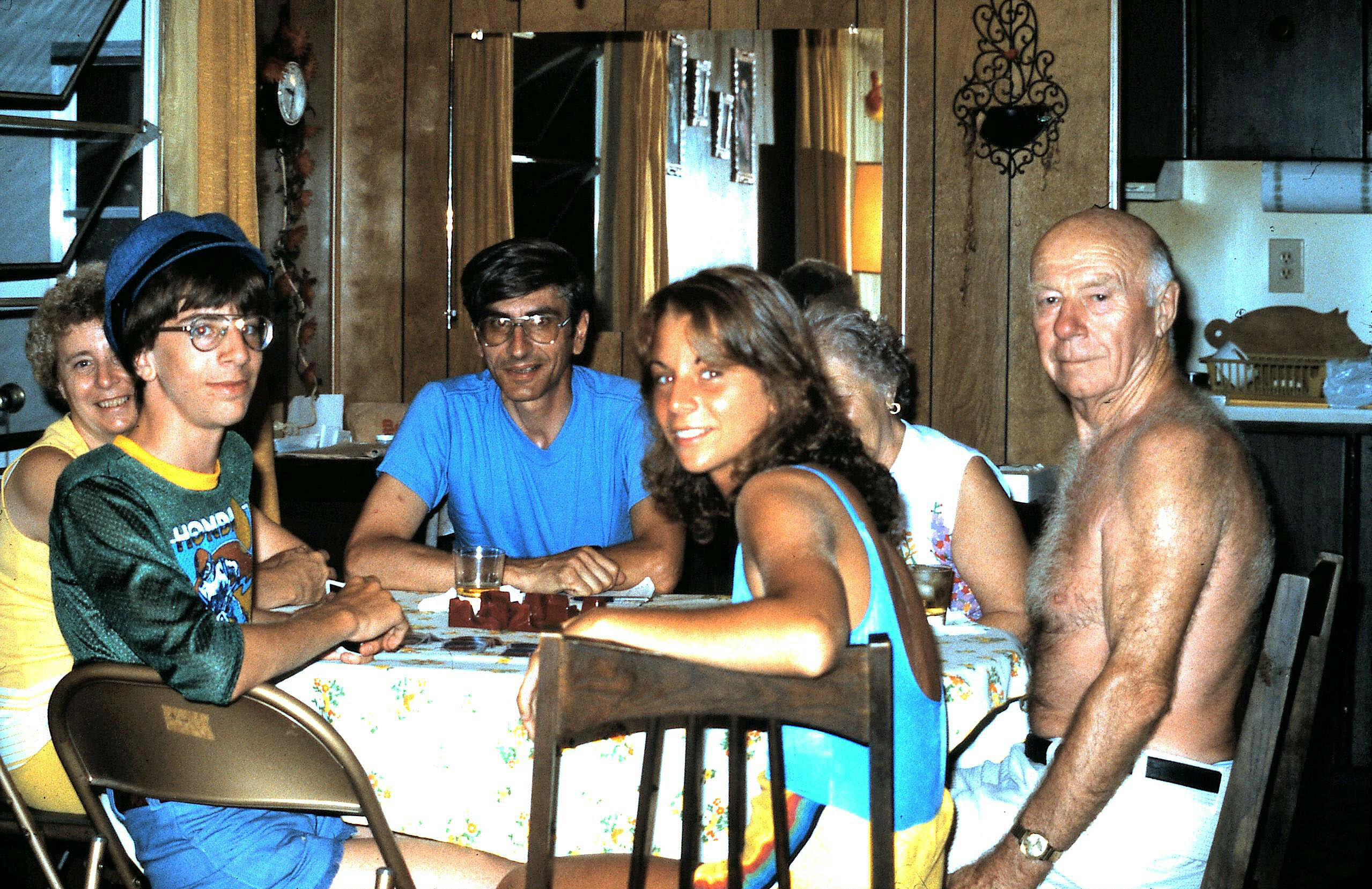 Group of people seated around a table playing a game, drinks on the table, one man shirtless on the right.