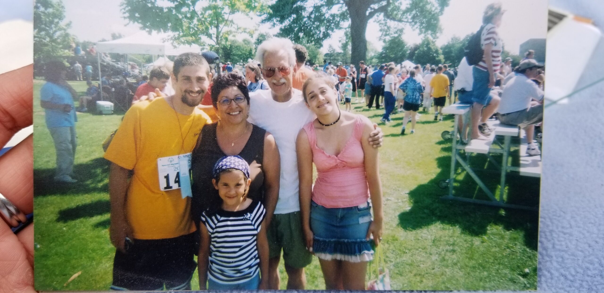 Five people posing together at a sunny outdoor park festival, smiling with a crowd and tents in the background.