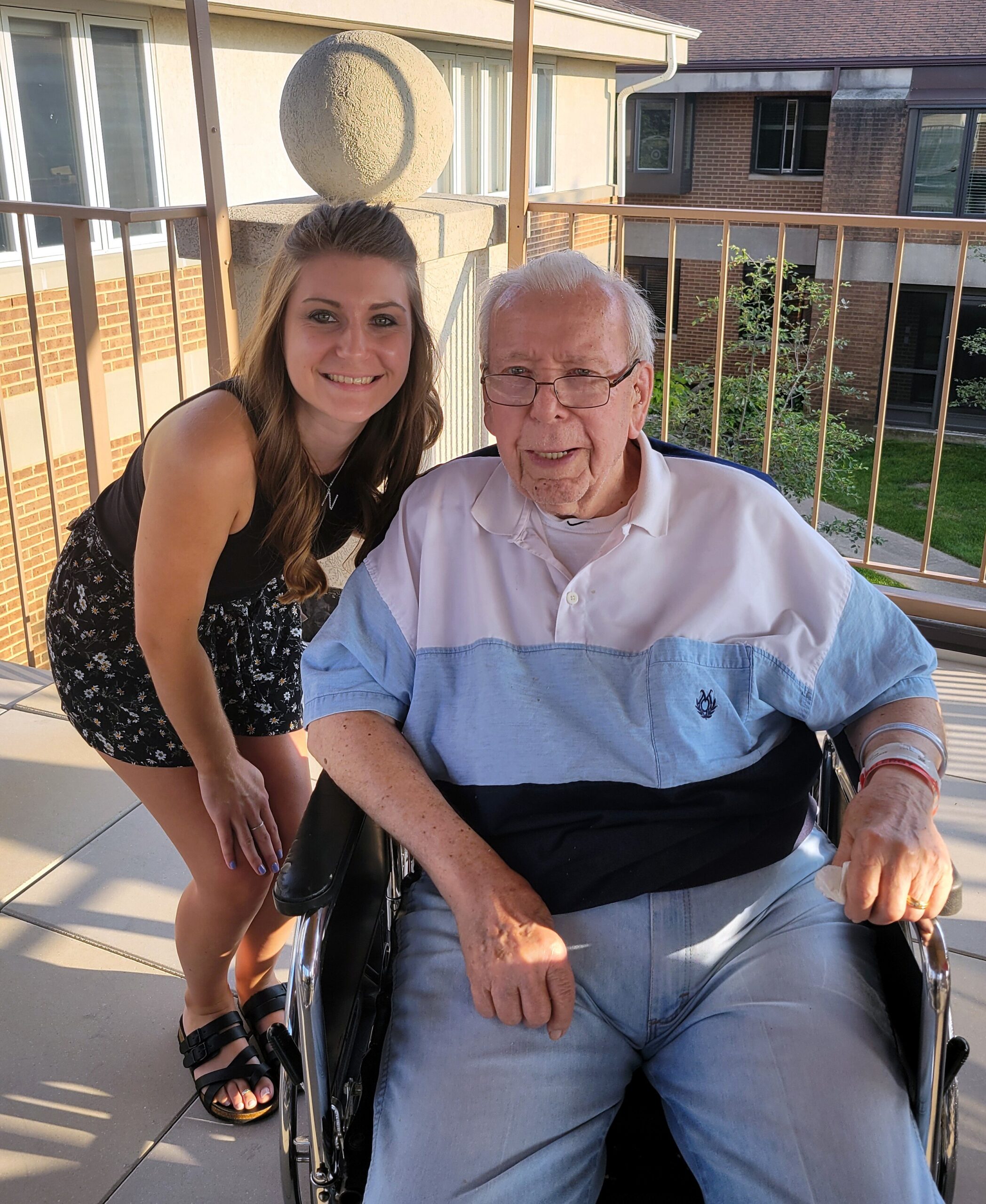 Young woman leaning beside a smiling elderly man in a wheelchair on a sunny balcony.