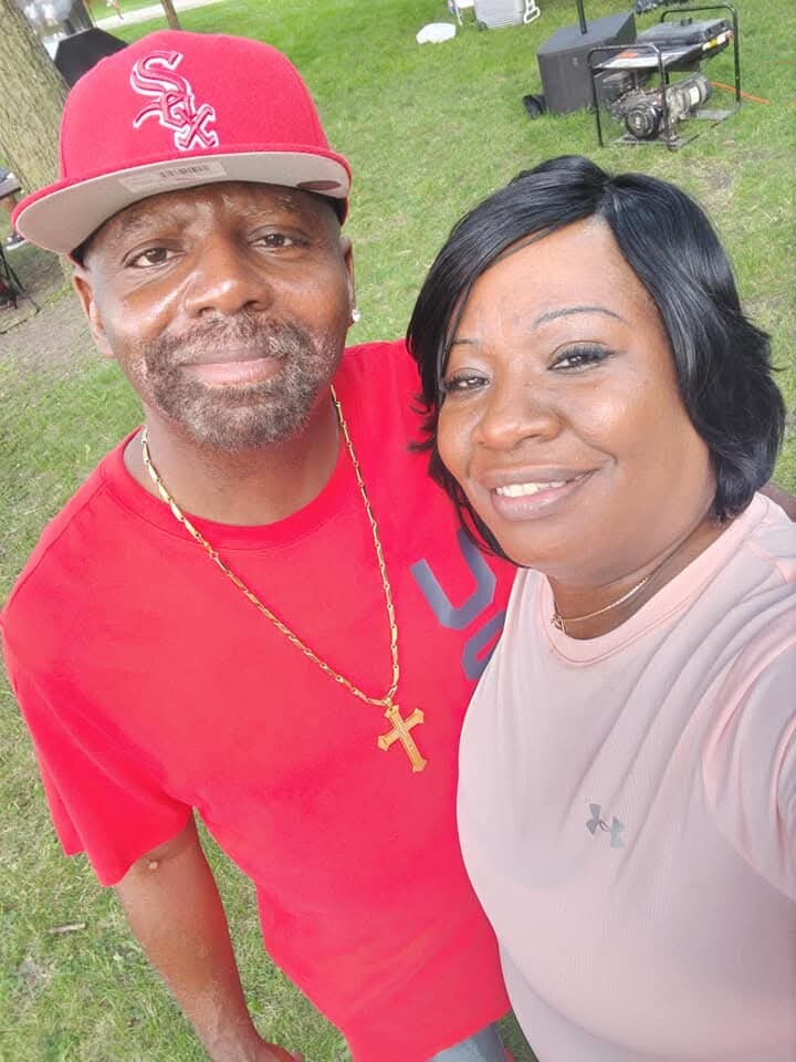 Smiling couple taking a selfie outdoors on grass; man in a red shirt and cap with a gold cross necklace, woman in a pale pink top nearby.