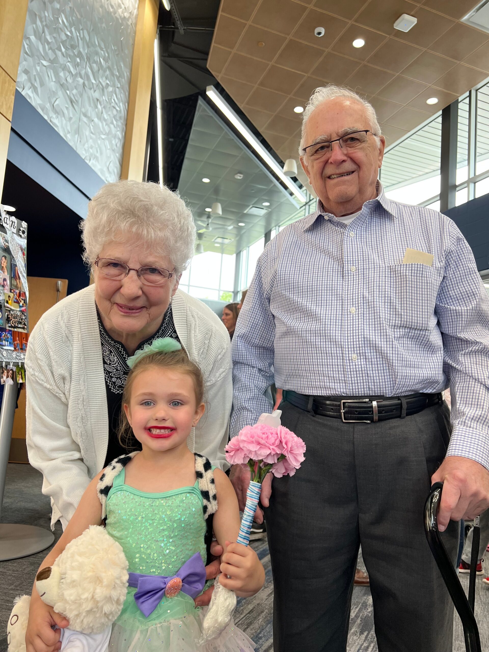 Three generations pose for a photo indoors: an elderly woman and man stand beside a smiling young girl in a turquoise dress holding a stuffed bear and a pink flower wand.