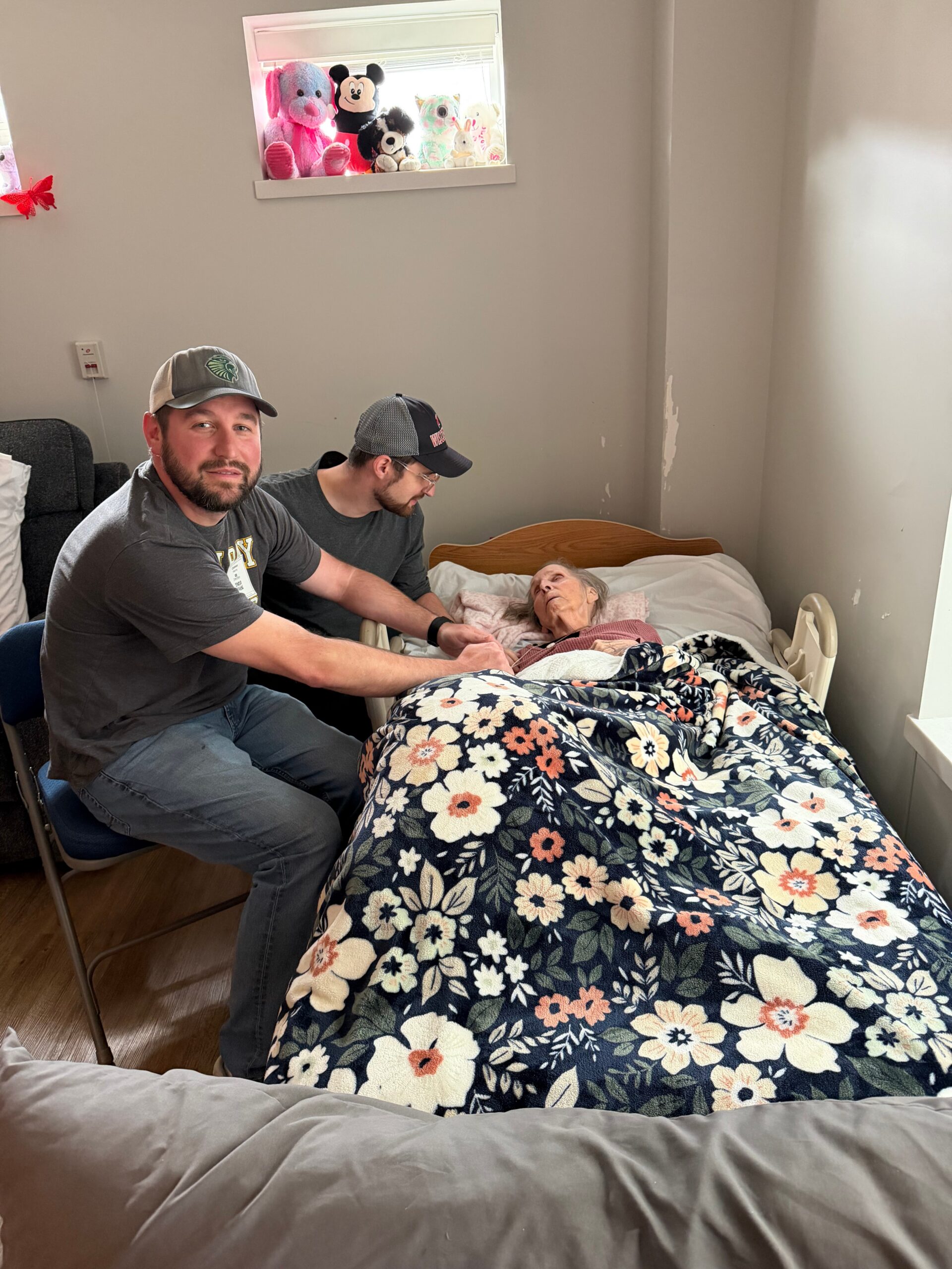 Two men sit by an elderly woman in bed in a small bedroom, a floral blanket covering her.