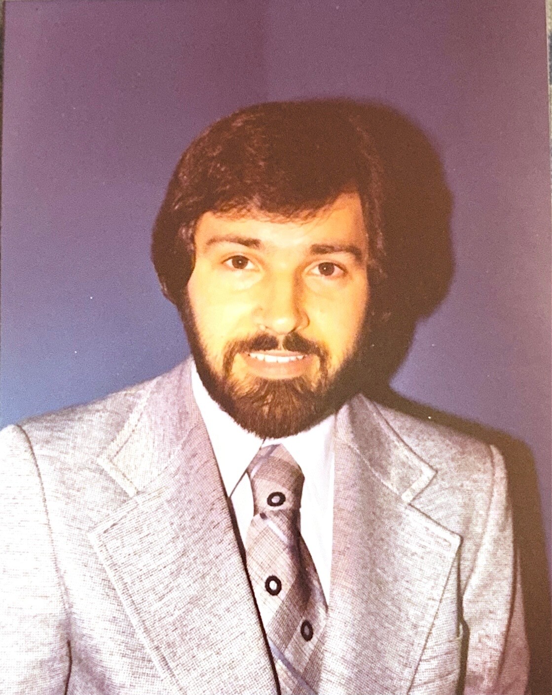 Portrait of a man with dark hair and a beard wearing a light gray suit and patterned tie against a purple backdrop