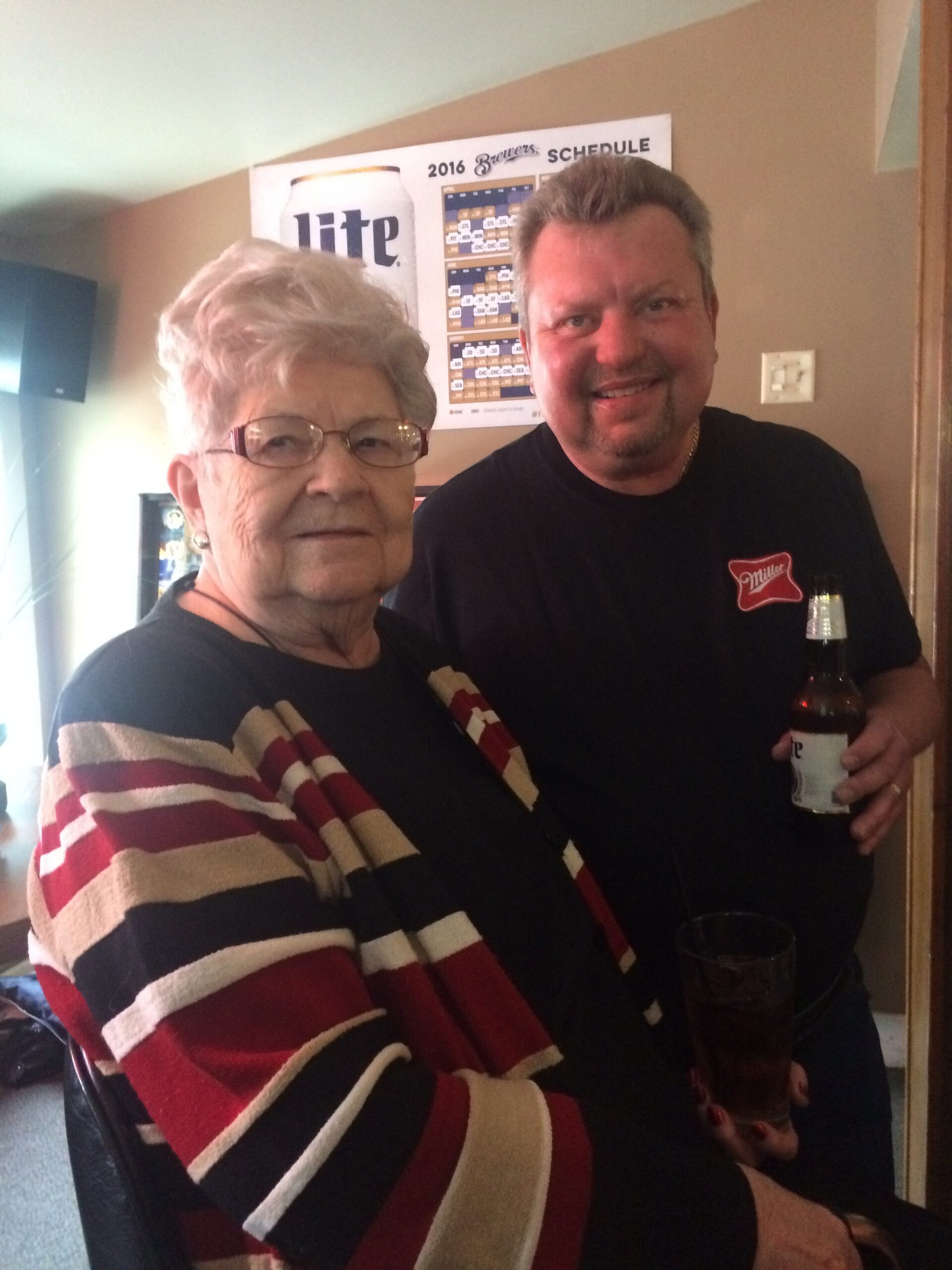 Older woman in a striped sweater poses with a smiling man holding a beer bottle in a room with a Brewers poster on the wall behind them.
