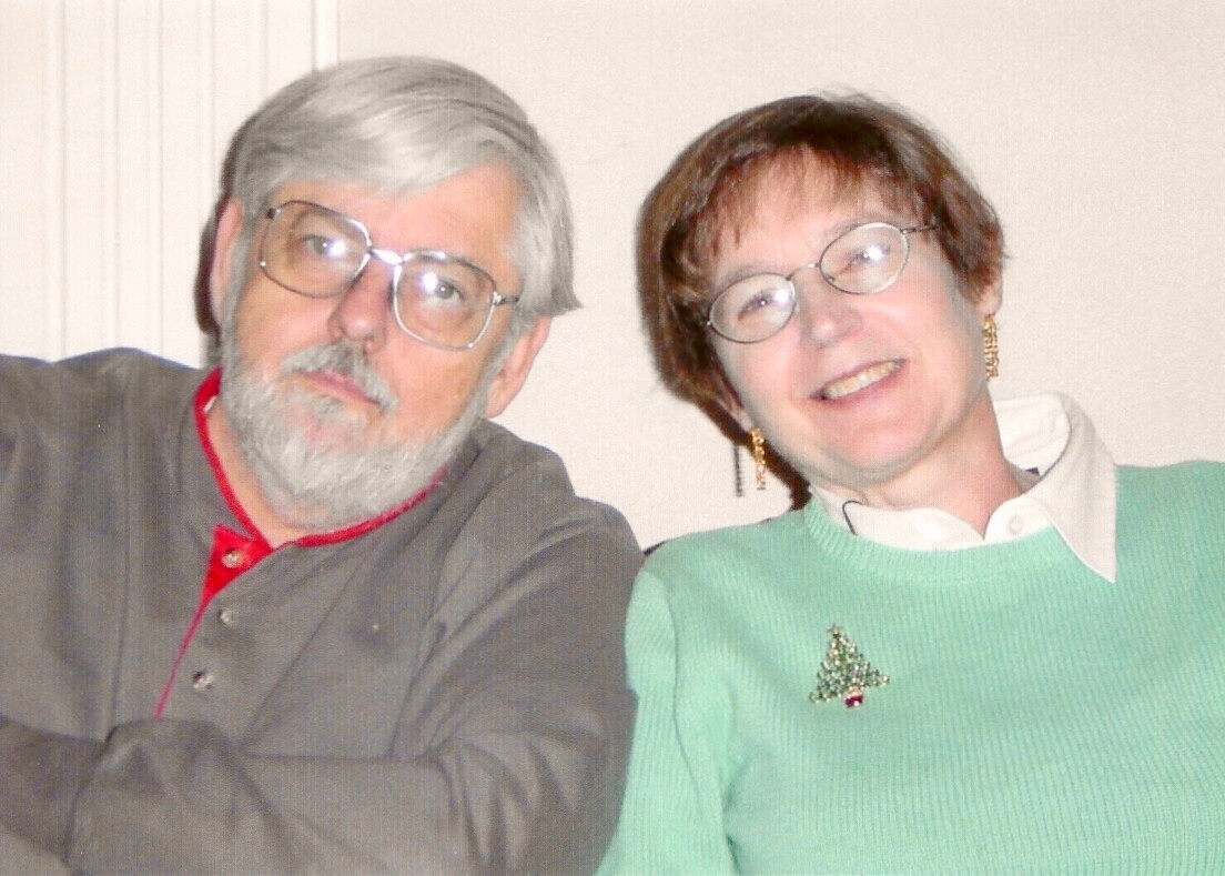 Two older adults leaning close together and smiling at the camera; man with gray hair and beard wearing glasses, woman with short hair in a green sweater with a Christmas tree pin.