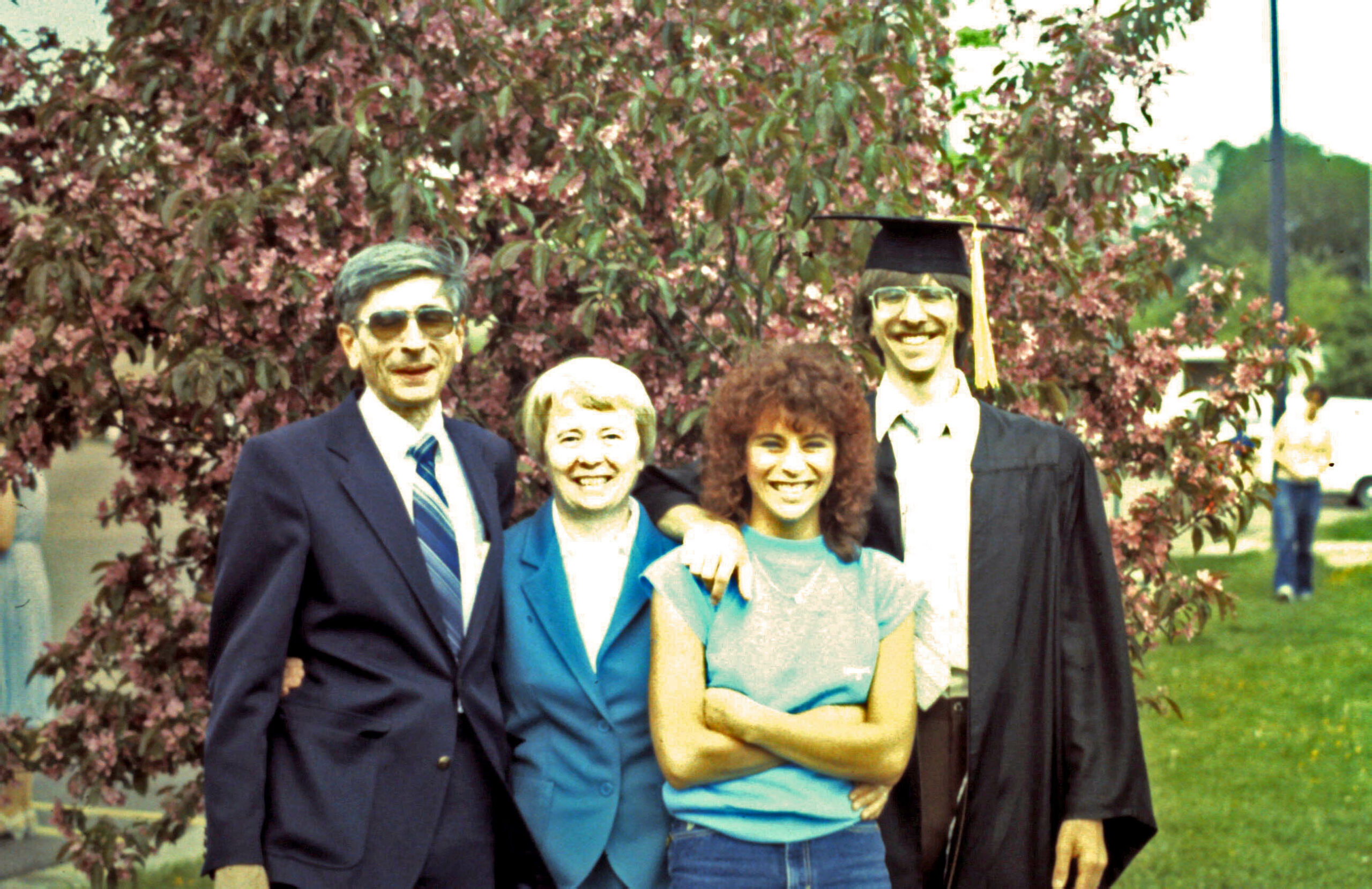 Group of five people posing outdoors, including a graduate in cap and gown, with smiling friends and a flowering tree in the background.