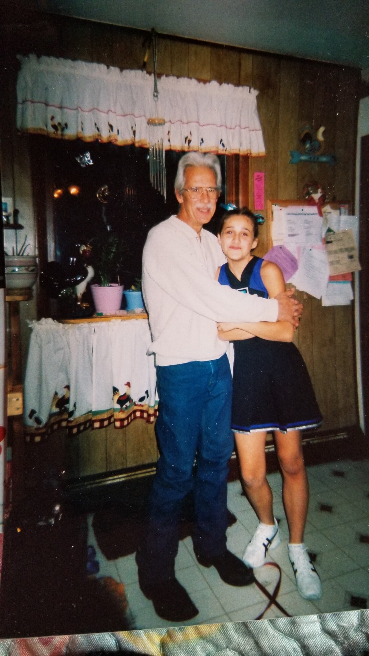 Grandfather with white hair and glasses hugs a girl in a blue basketball jersey in a cozy kitchen setting.