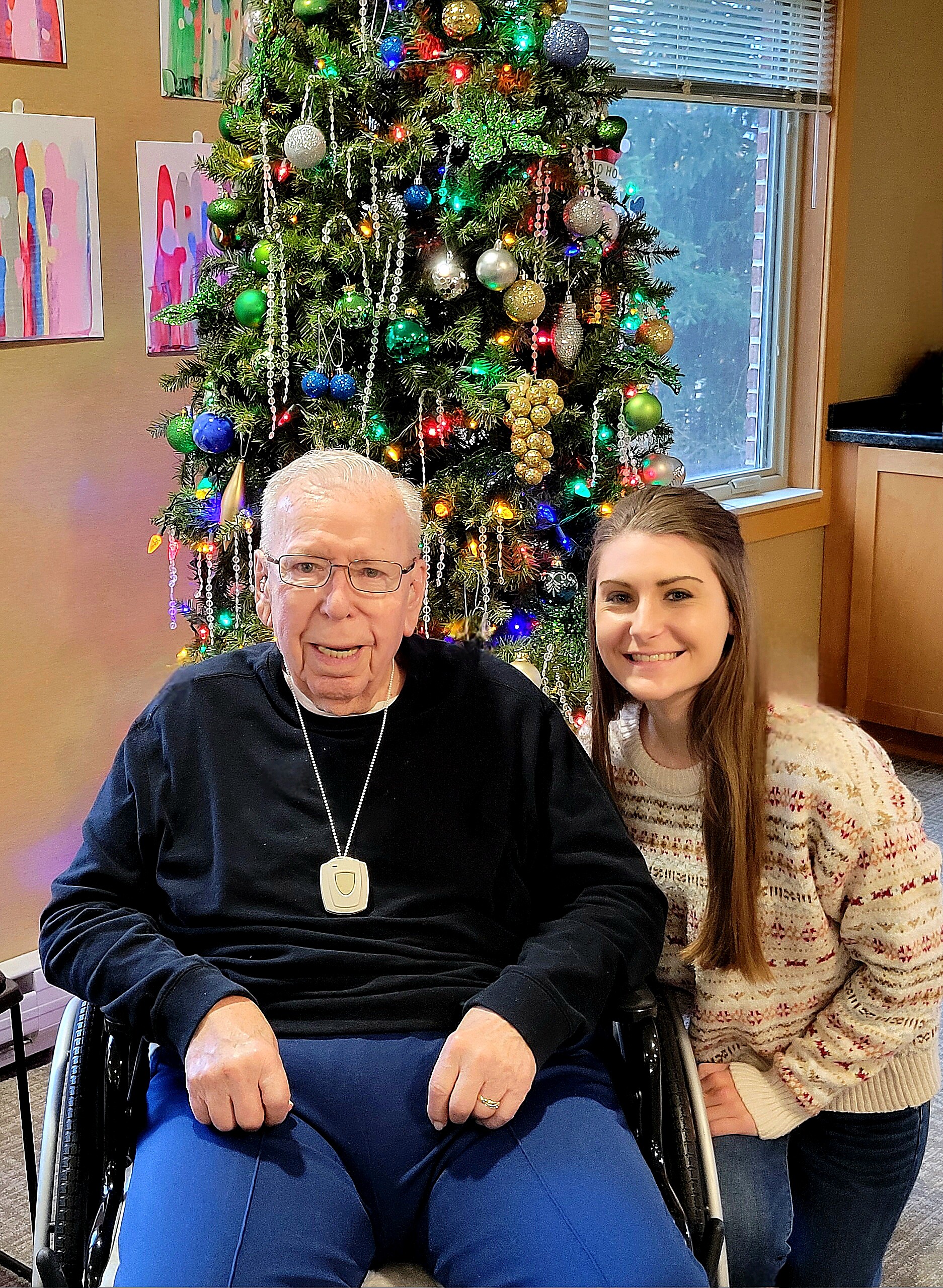 Man in a wheelchair and a young woman smiling beside a decorated Christmas tree indoors.