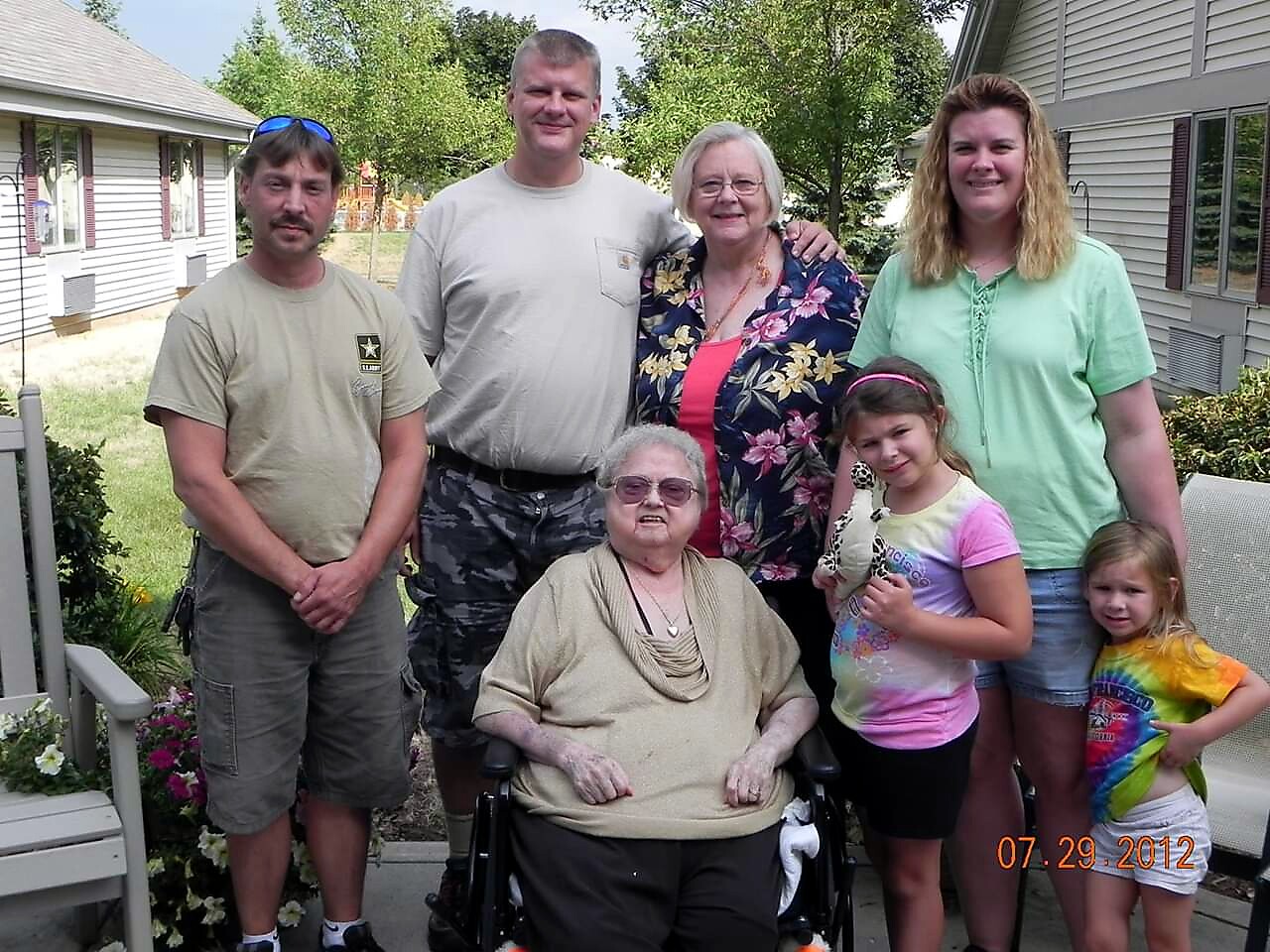Seven family members pose outdoors in a backyard, with an elderly woman in a wheelchair at the center, all smiling. Date stamp reads 07.29.2012 on the bottom corner.