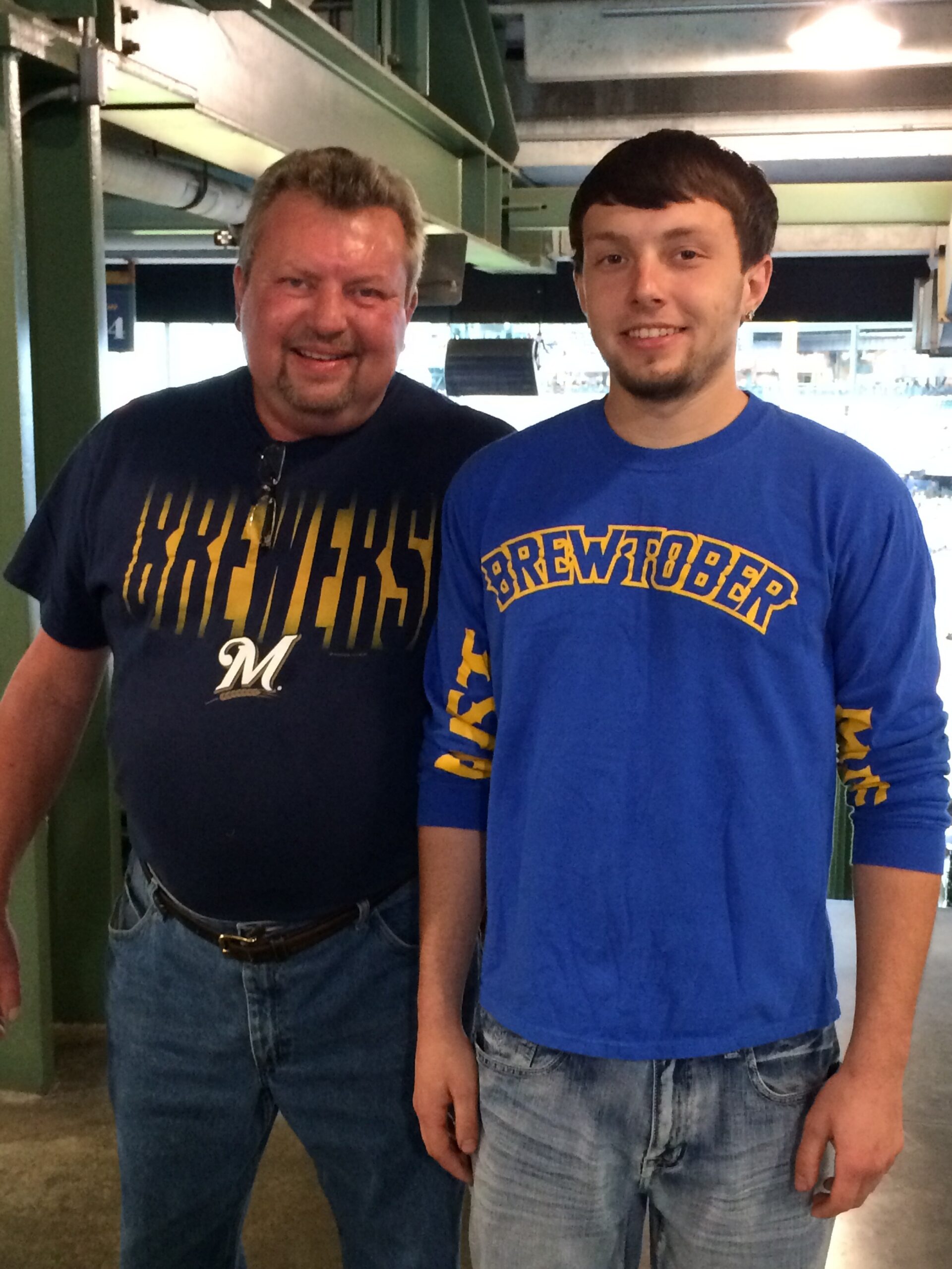 Two men posing for a photo in an industrial setting, both smiling and wearing Brewers-branded shirts (blue and navy).