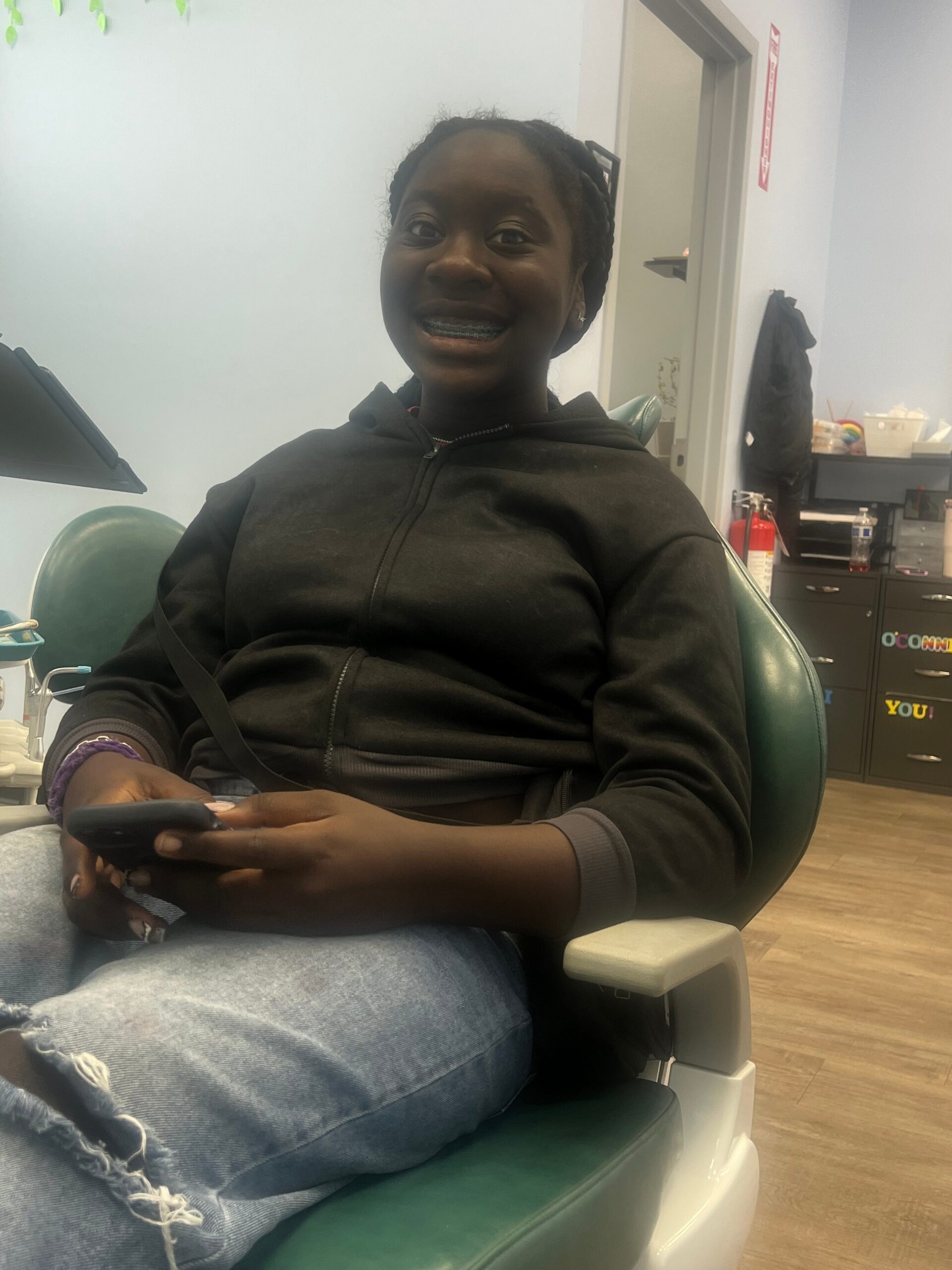 Girl wearing a black hoodie and jeans sits in a dental chair, smiling with braces, holding a phone in her lap in a clinic setting.