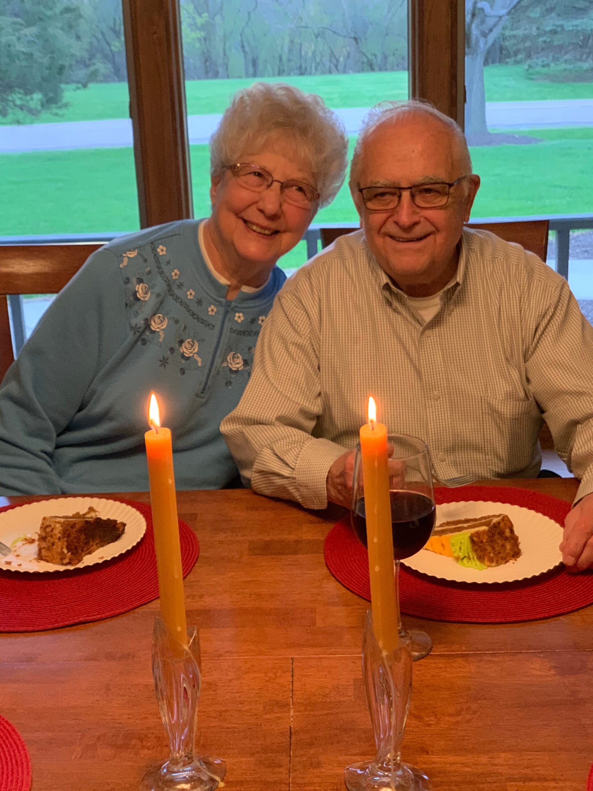 Smiling elderly couple at a dining table with two lit candles, cake slices, and a glass of red wine.