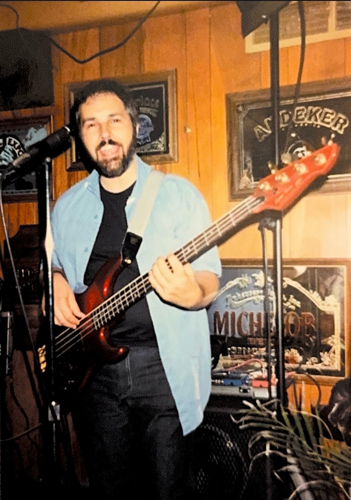 Male musician playing a red bass guitar on a small stage in a bar, wearing a light blue shirt over a black tee with a microphone nearby.