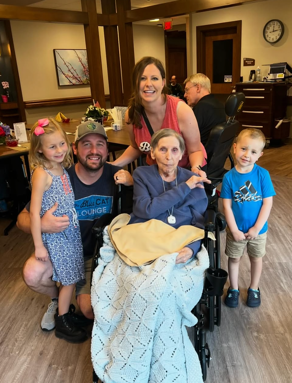 Five family members pose with an elderly woman in a wheelchair in a lobby-like room, all smiling at the camera.