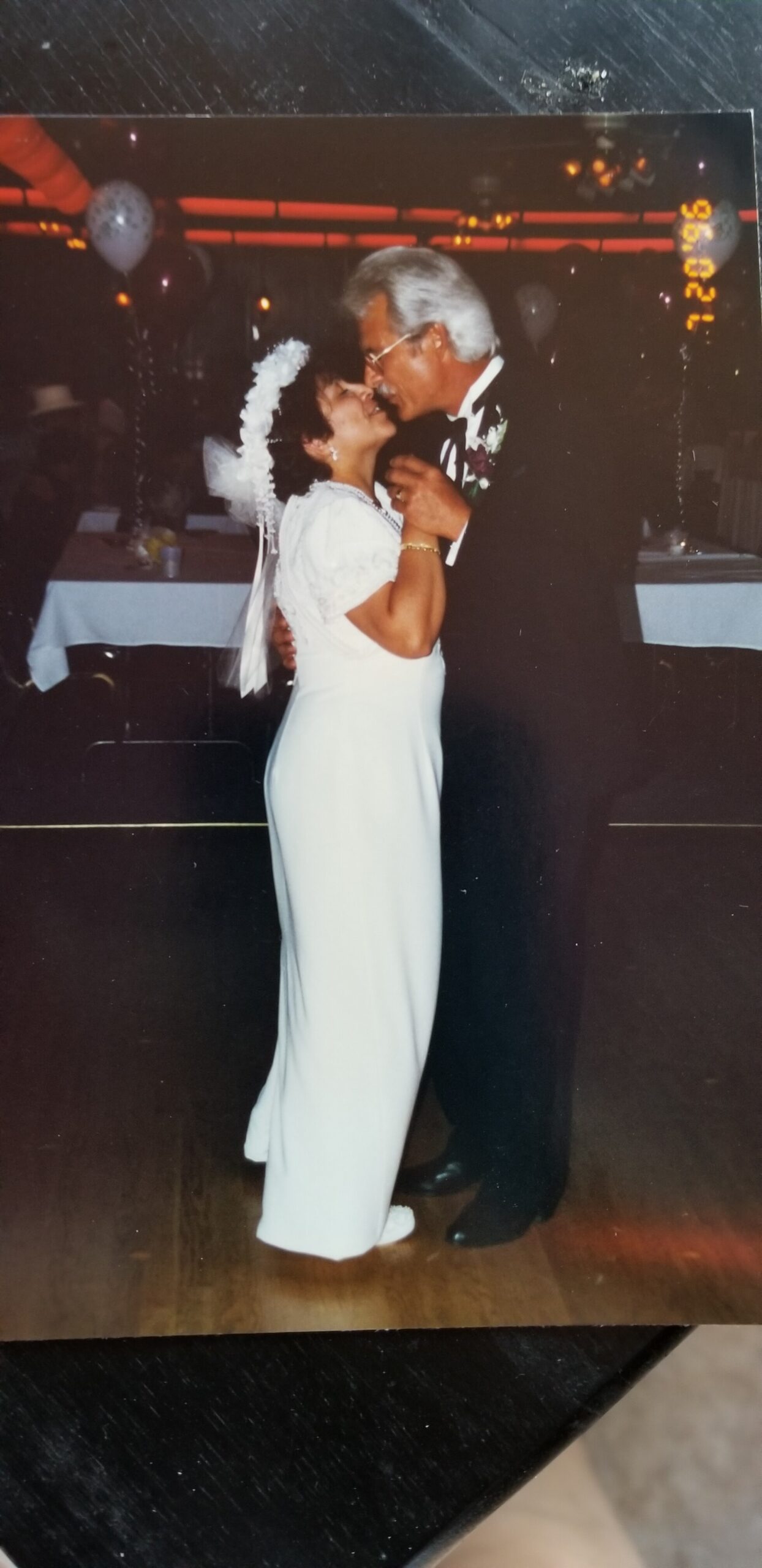 Older couple dancing closely at a wedding reception; the bride in a white dress with veil and the groom in a suit smiling at each other.