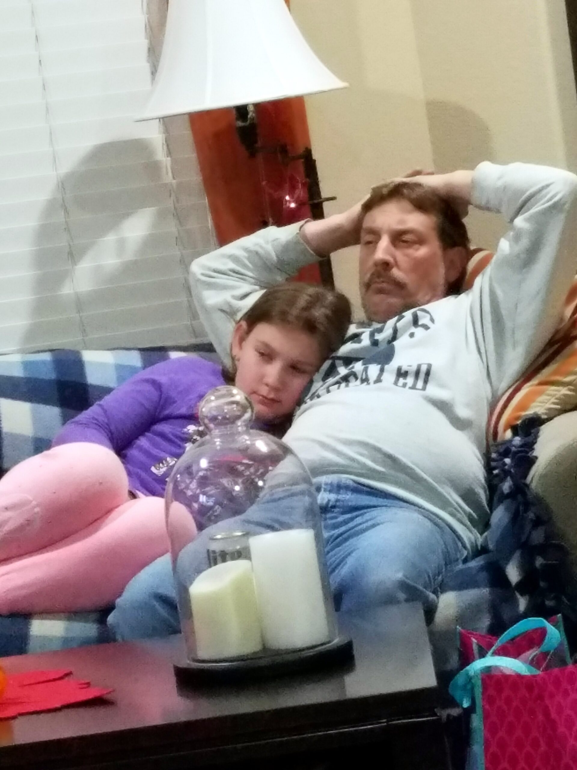 Man with a mustache relaxing on a couch with hands behind head, a child resting beside him, in a living room with candles on the table nearby.
