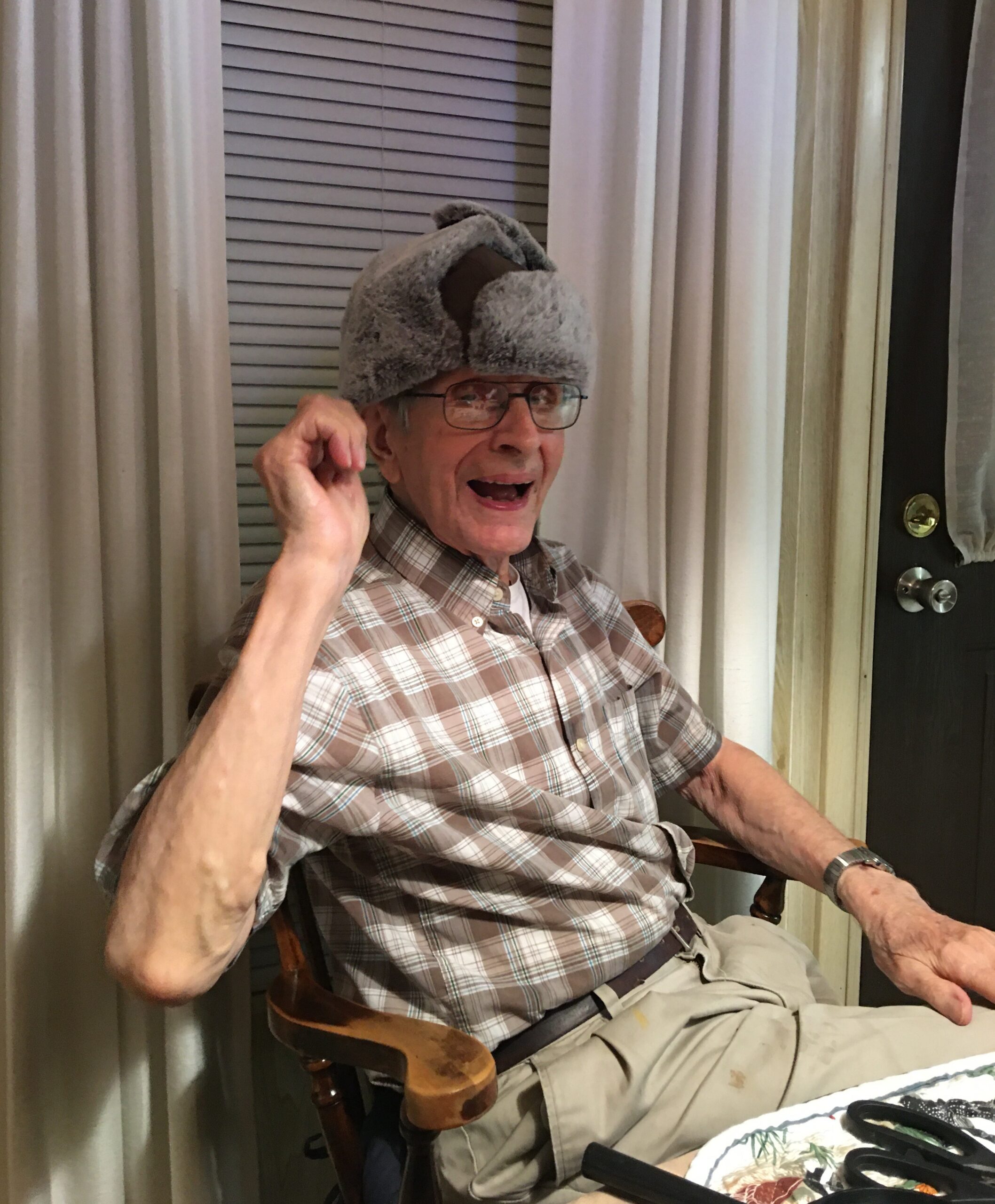 Older man wearing a fluffy gray hat sits in a wooden chair, smiling and laughing indoors beside beige curtains and a door.