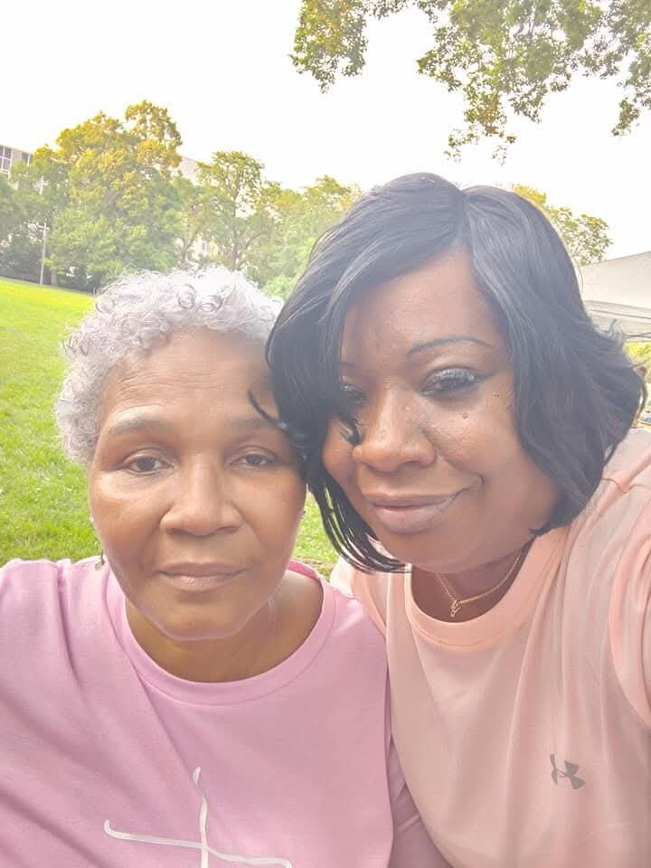 Two women posing for a selfie outdoors on a sunny day, with green grass and trees in the background.