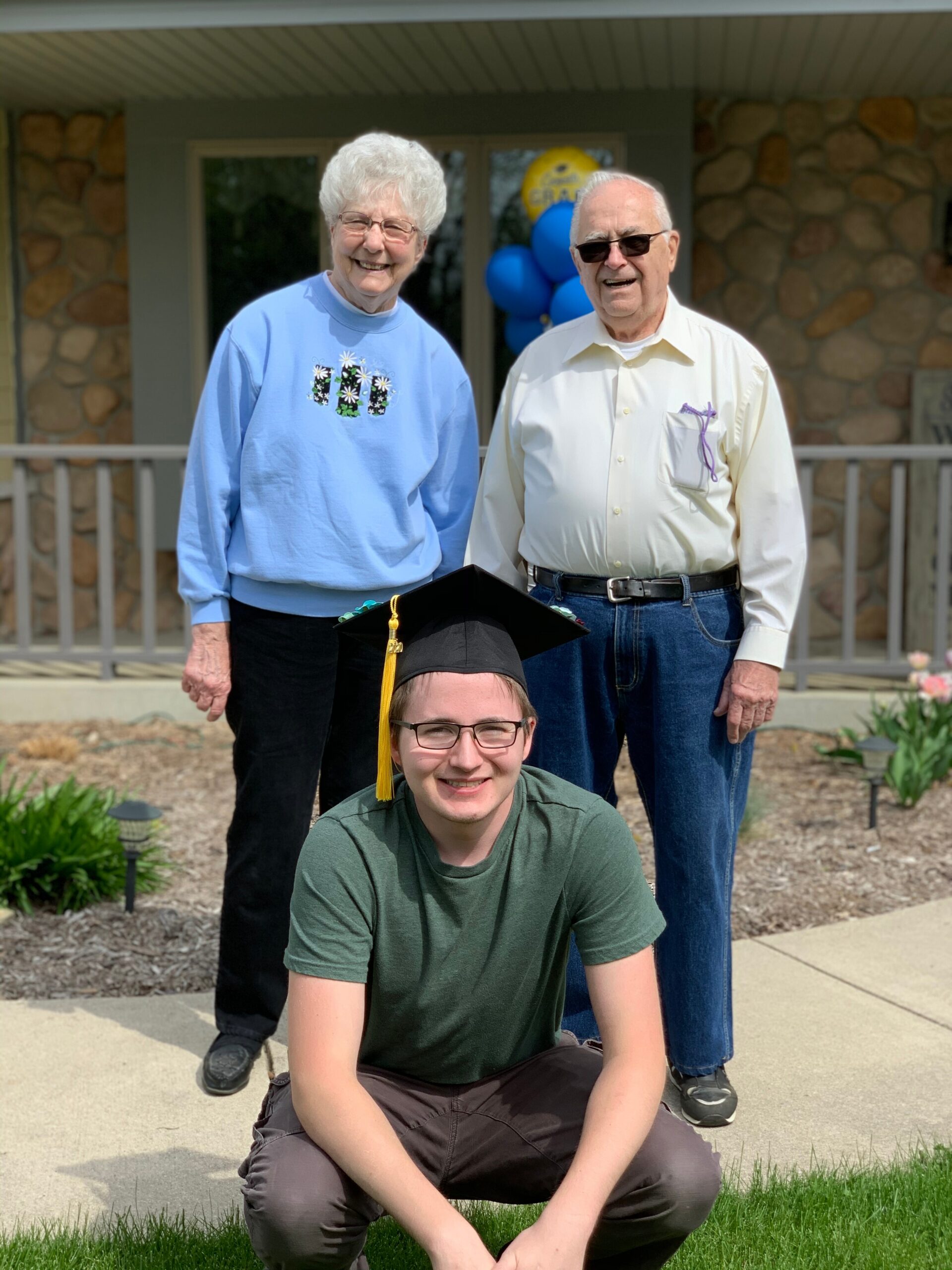 Graduating student in front wearing cap with tassel, flanked by smiling elderly couple on a porch with balloons in the background.