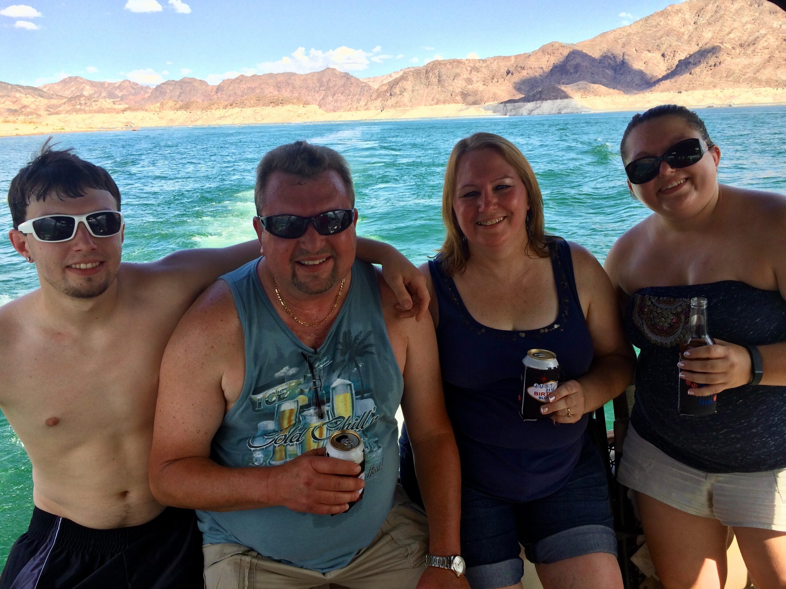 Group of four adults smiling on a boat, holding drinks, with turquoise water and rocky desert mountains in the background