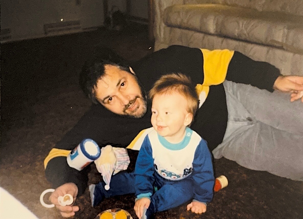 Man with beard lying on the floor next to a smiling toddler playing with toys in a living room.