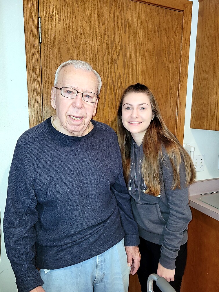 Smiling elderly man in a navy sweater and a younger woman in a gray hoodie pose together in a home kitchen with wooden cabinets behind them.