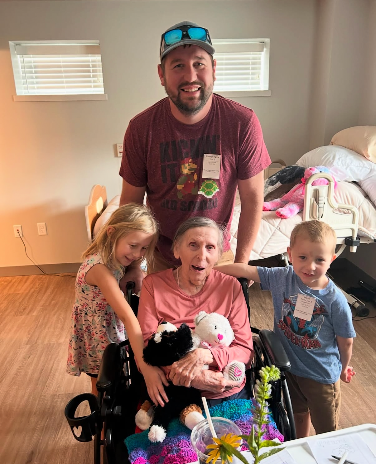 Man standing behind a smiling elderly woman in a wheelchair, with a girl and a boy beside her in a cozy room full of toys and blankets.