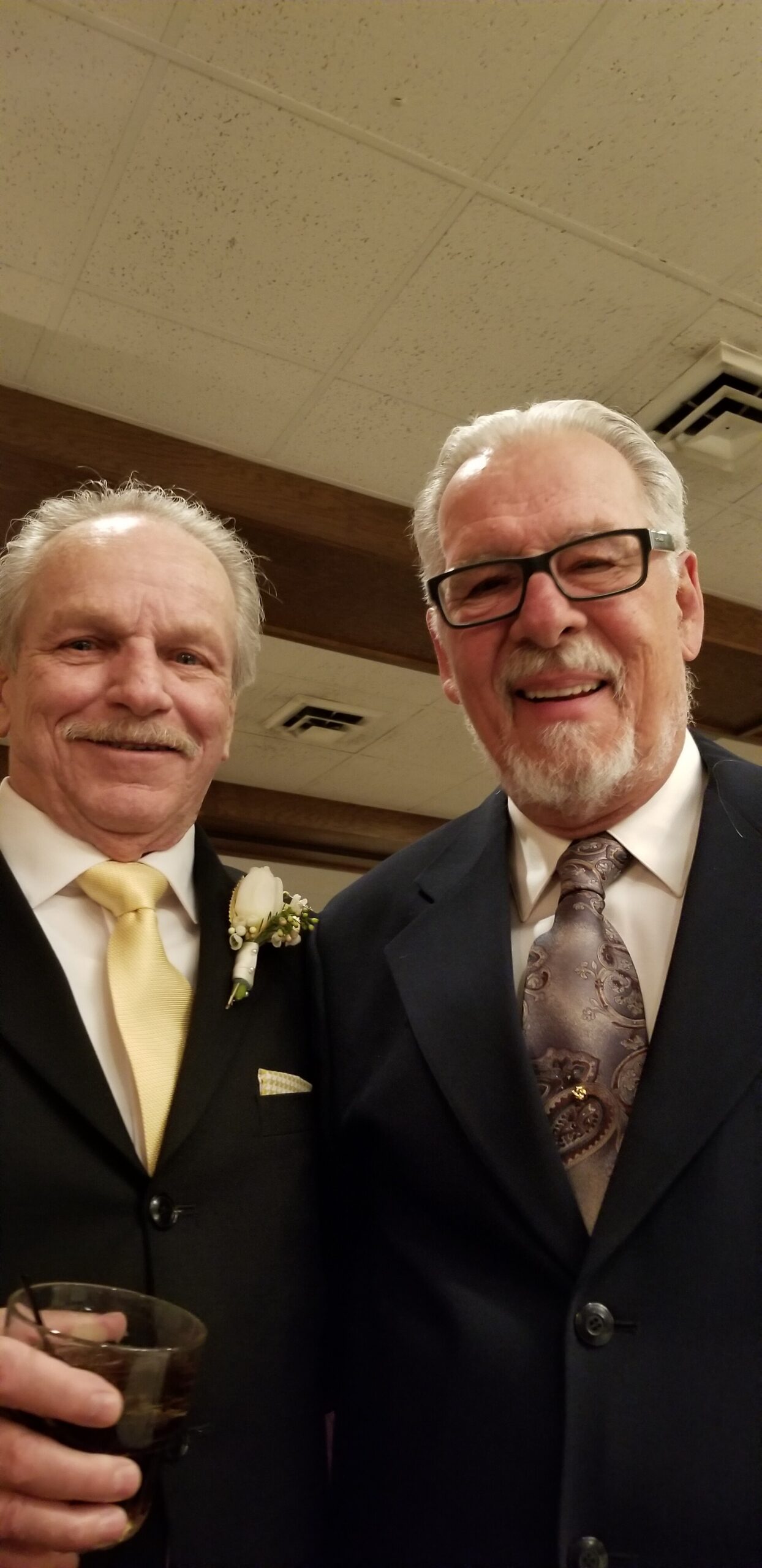 Two older men in suits posing for a selfie at a formal event; boutonniere on left, yellow tie, drink in hand visible below.