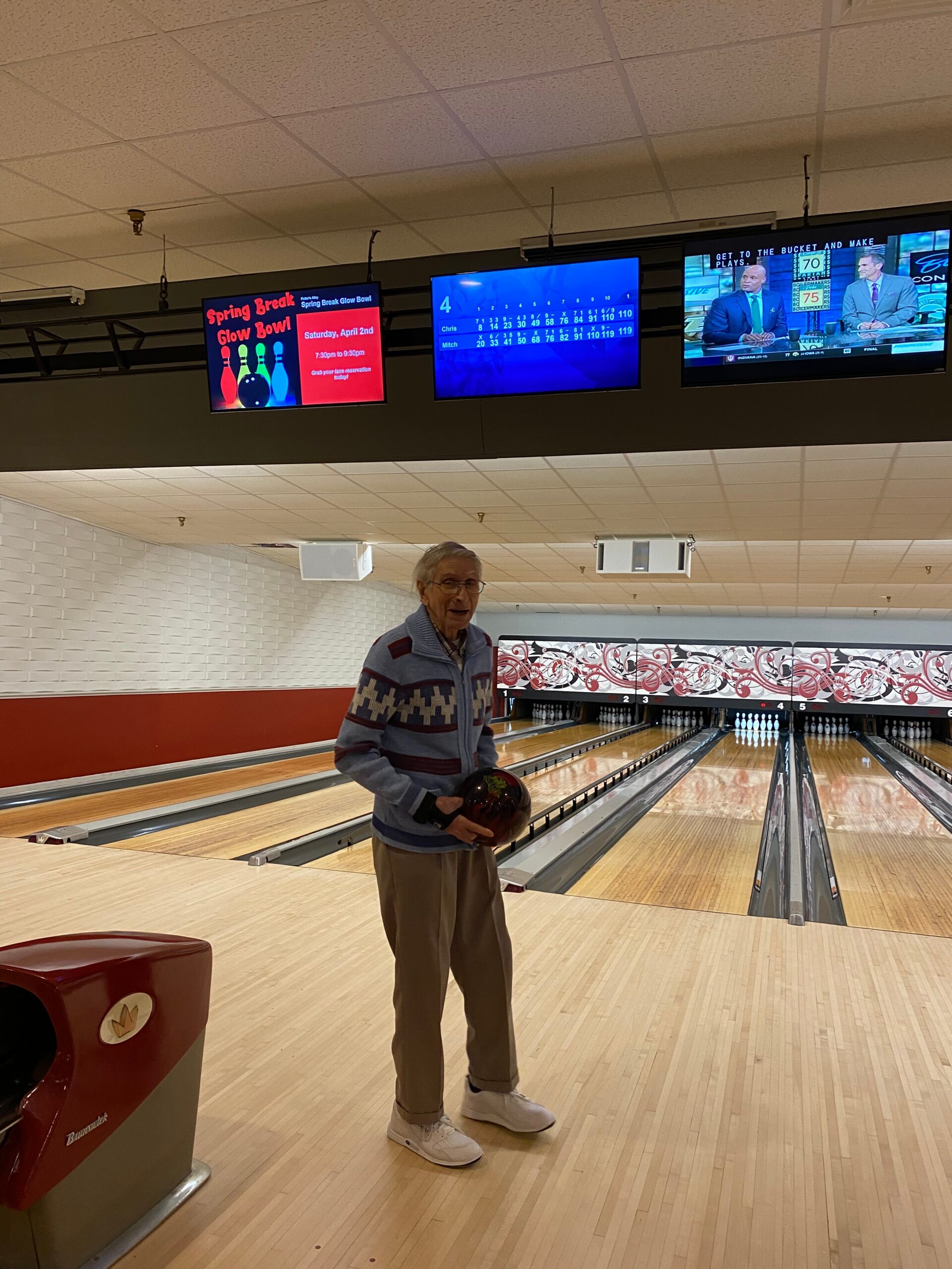 Elderly man in a striped sweater stands on a bowling lane, holding a red bowling ball, ready to bowl.