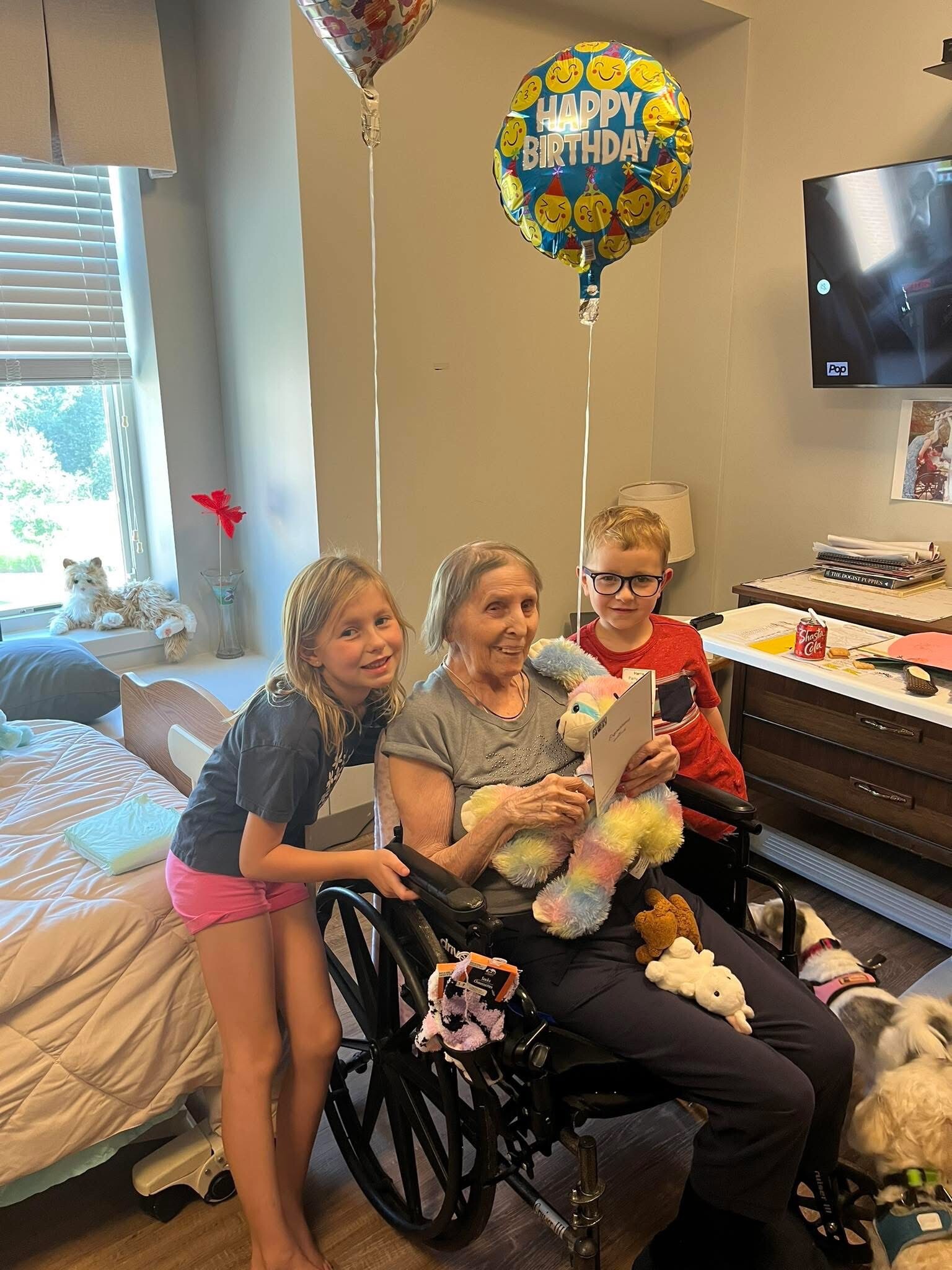 Two children hug an elderly woman in a wheelchair during a birthday celebration with bright balloons and toys nearby.