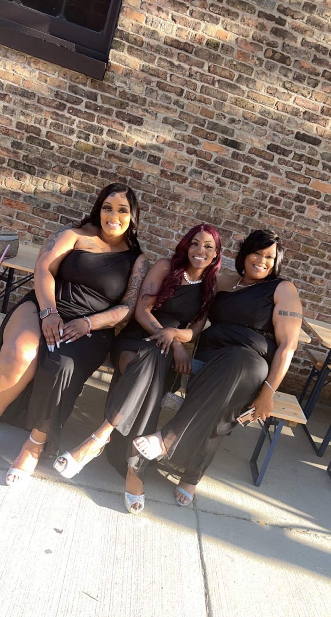 Three women in black dresses sit on a bench against a brick wall, smiling at the camera.