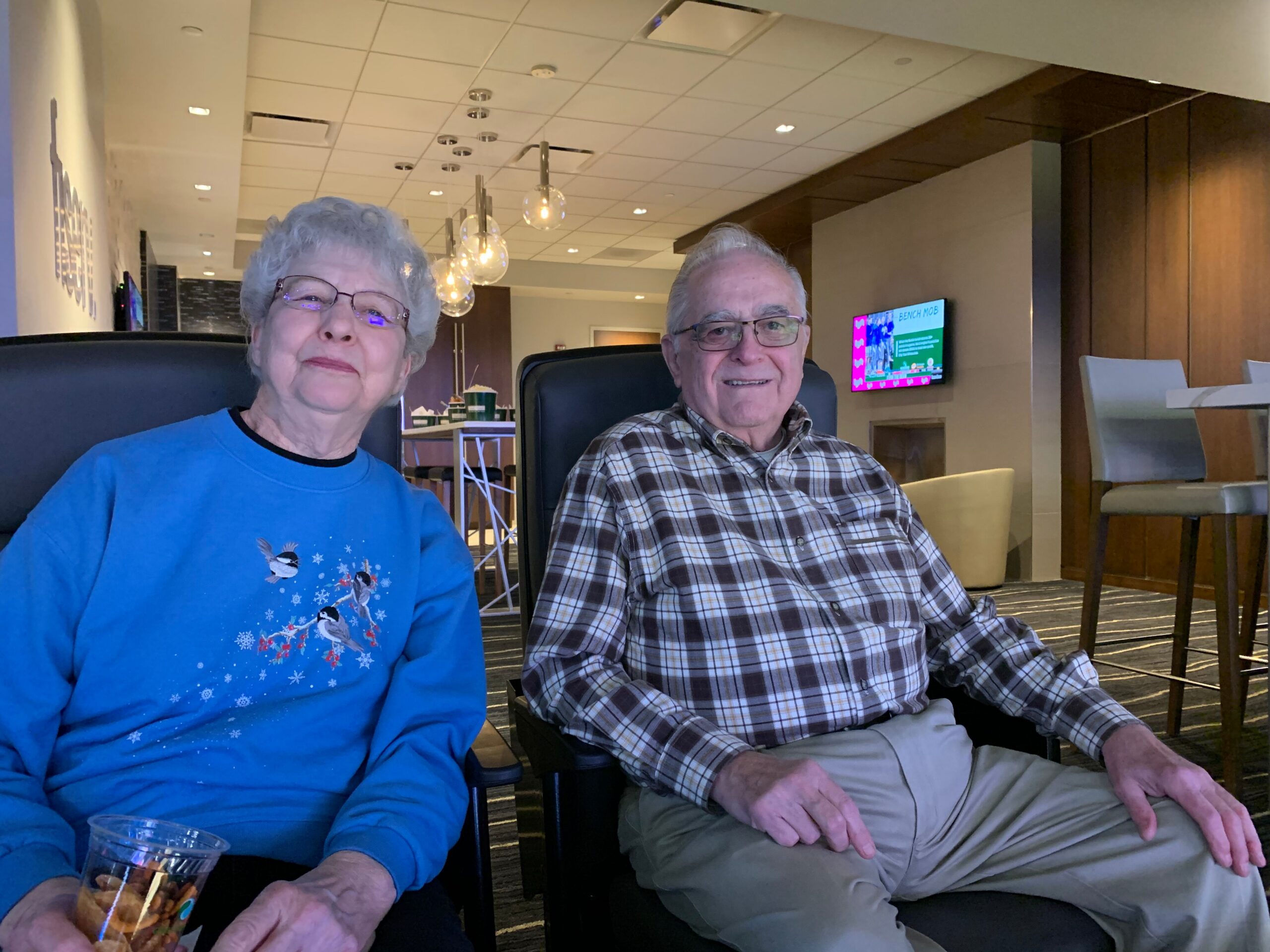 Two elderly people seated in a modern lounge, smiling at the camera; woman in a blue sweater on the left and man in a plaid shirt on the right.