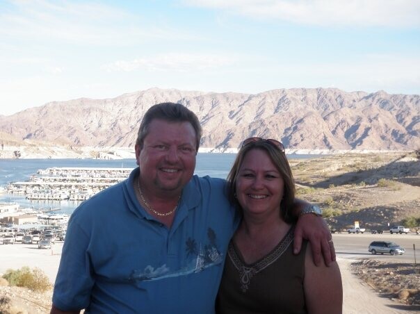 Smiling couple stands together with their arms around each other, in front of a marina and rugged desert mountains under a blue sky.