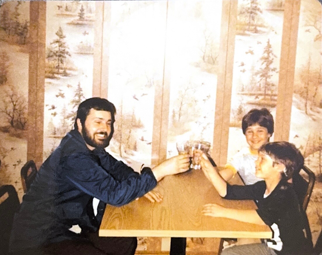 Man with a beard and two children clinking glasses over a wooden table, smiling at a indoors gathering.