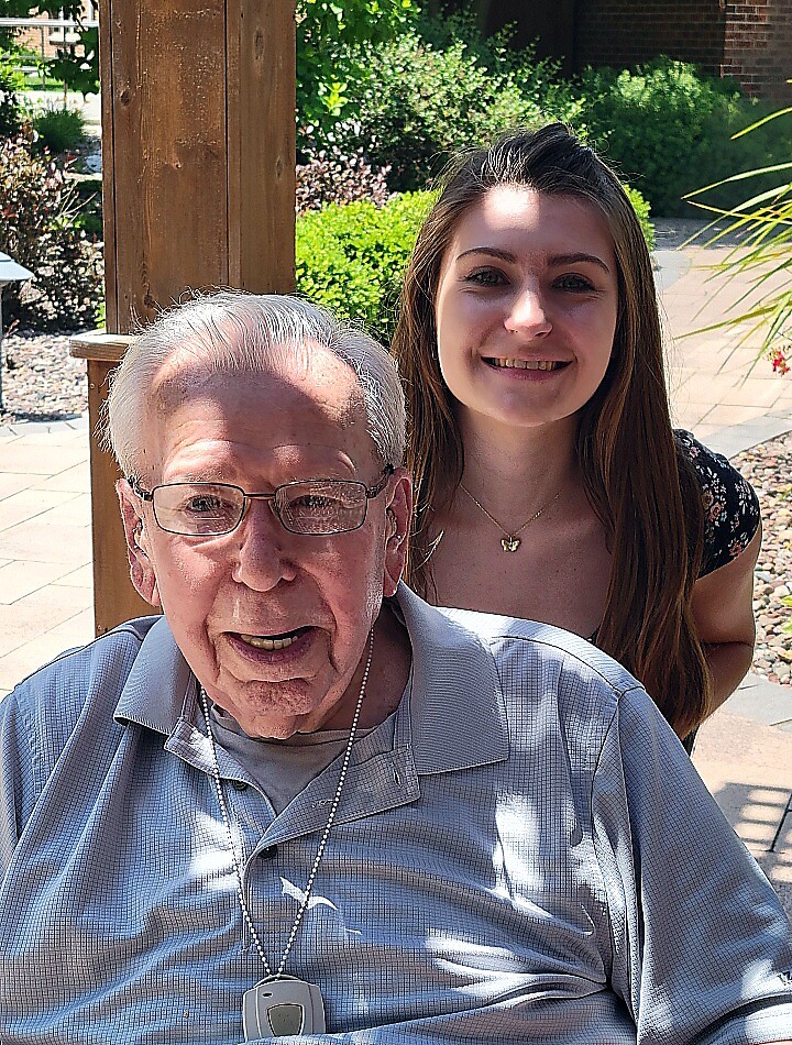 Smiling elderly man with glasses sits in the foreground, wearing a light gray polo shirt and a medical alert pendant; a smiling young woman stands behind him outdoors in a sunny garden setting.