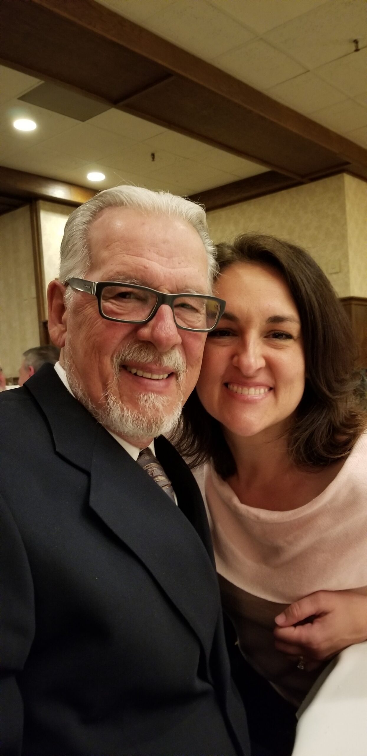 Smiling older man with gray hair and glasses in a dark suit posing for a selfie with a smiling woman in a pink sweater.