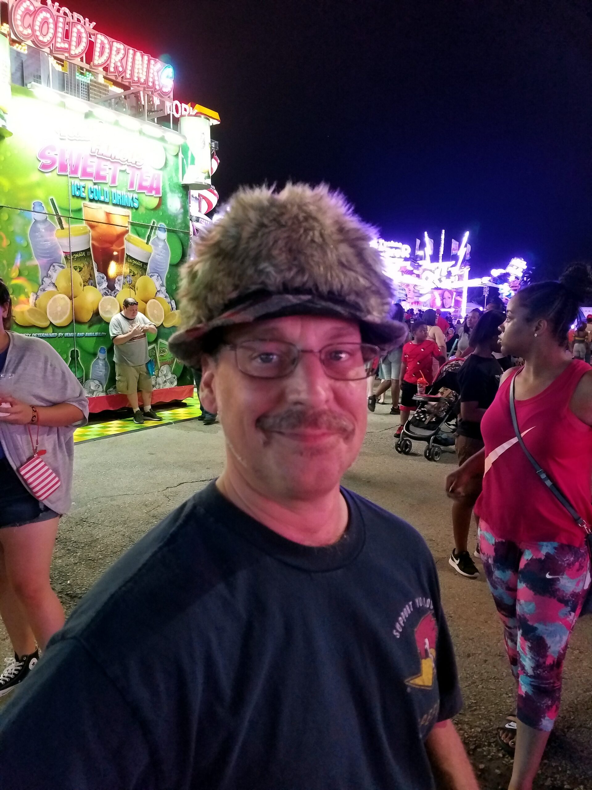 Man wearing a furry hat takes a selfie at a crowded nighttime carnival with bright, colorful signs and rides in the background, including a lemonade/tea stand.