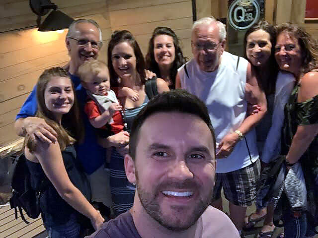 Smiling man in foreground taking a selfie with a large group of family and friends posing on a wooden deck, including a baby held by a woman.
