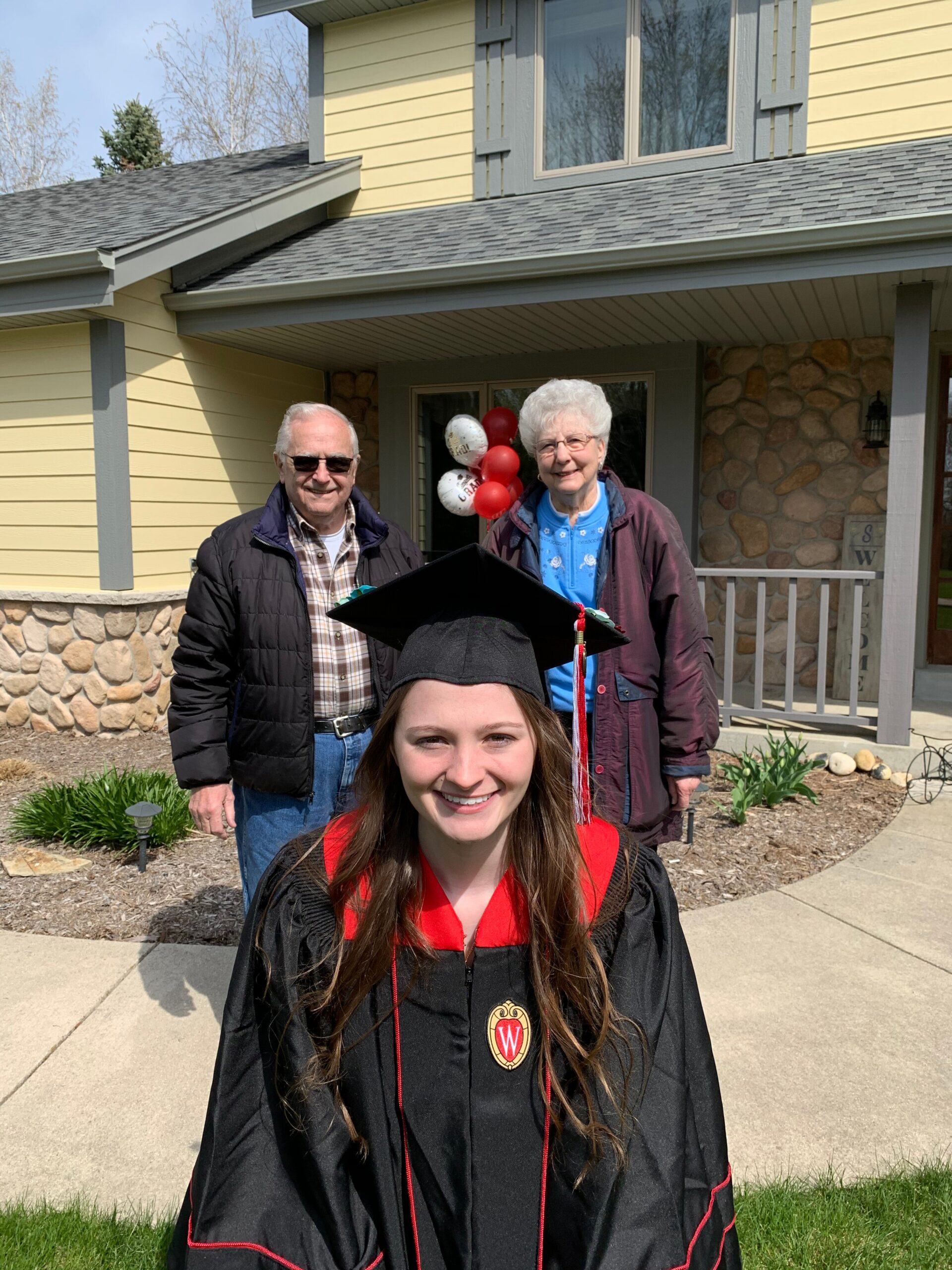 Smiling graduate in a black gown with red trim in the foreground, with an older couple and red balloons behind near a yellow house.