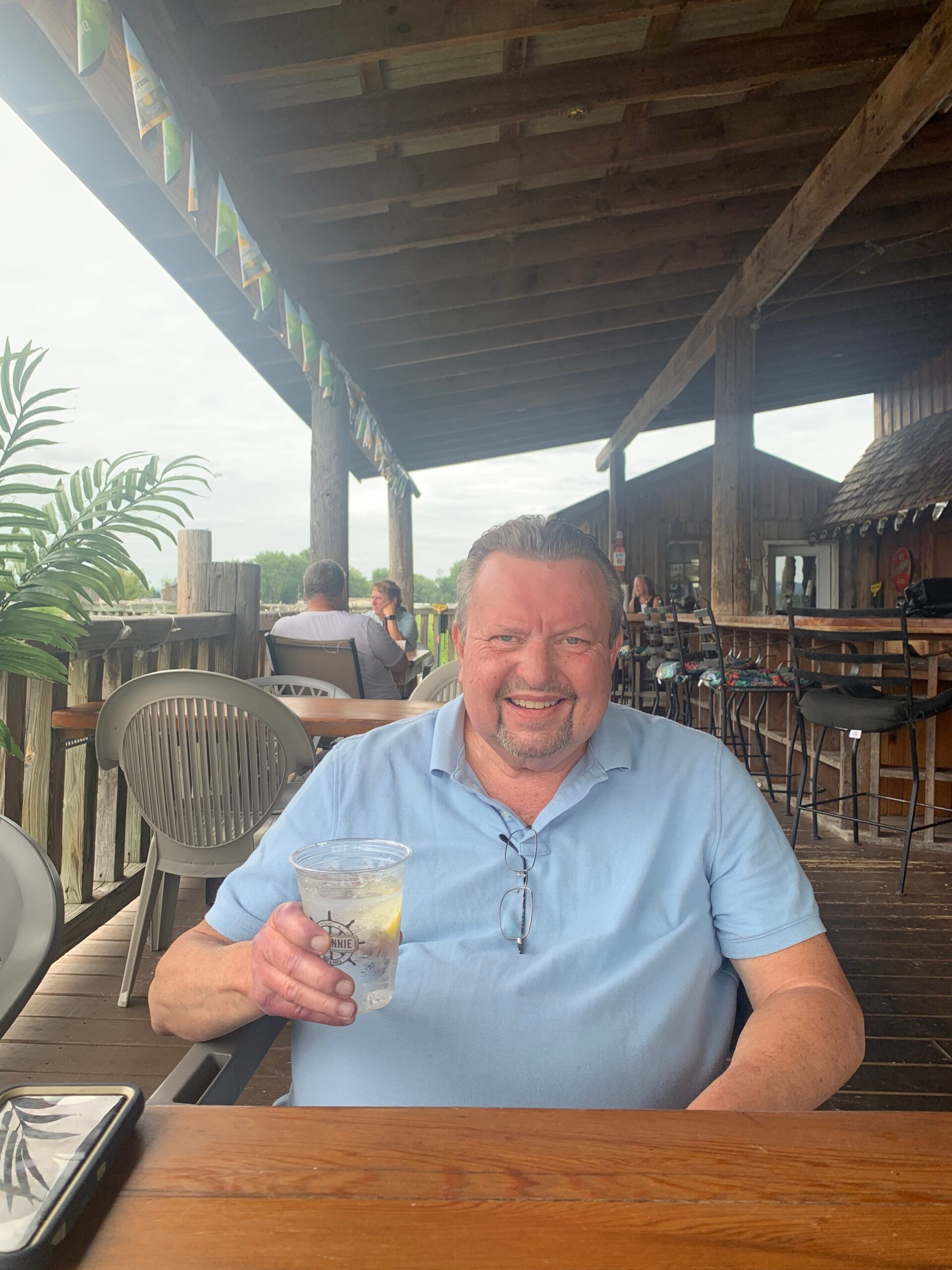 Smiling man in a light blue polo sitting at an outdoor wooden cafe table, holding a clear plastic cup with ice. Behind him are wooden posts, a shaded roof, and string of banners along the railing.