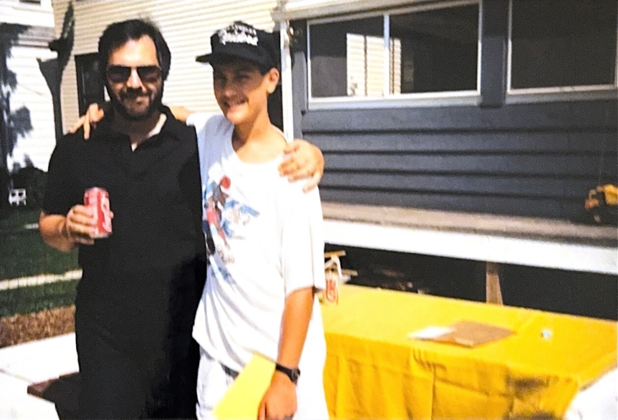 Two men pose together outside a house, one in a black shirt holding a Coca‑Cola can, the other in a white graphic tee and cap with his arm around his friend.