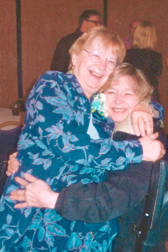 Two elderly women embrace and smile at a social gathering, one wearing a teal floral jacket.