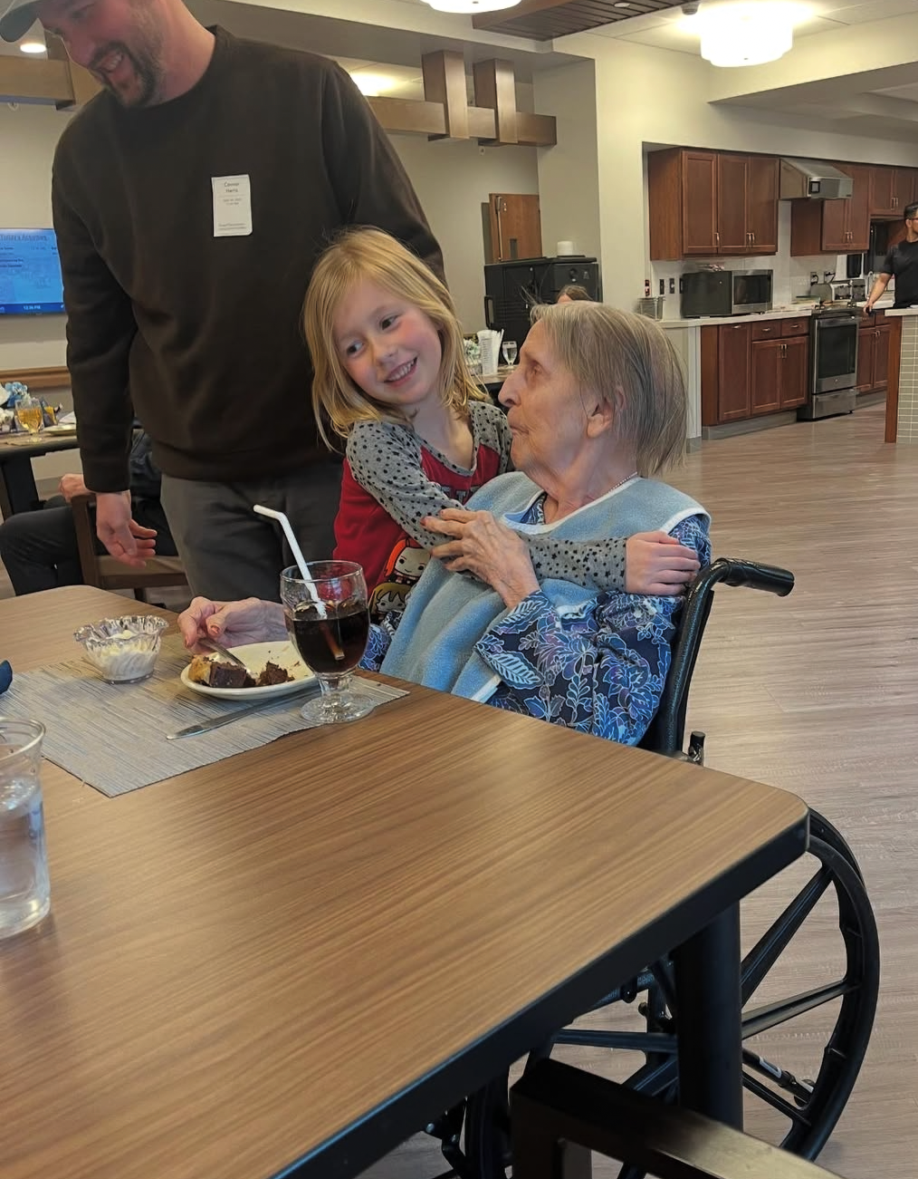 A young girl hugs an elderly woman in a wheelchair at a dining table, sharing a smile over a plate of cake and a glass of soda.