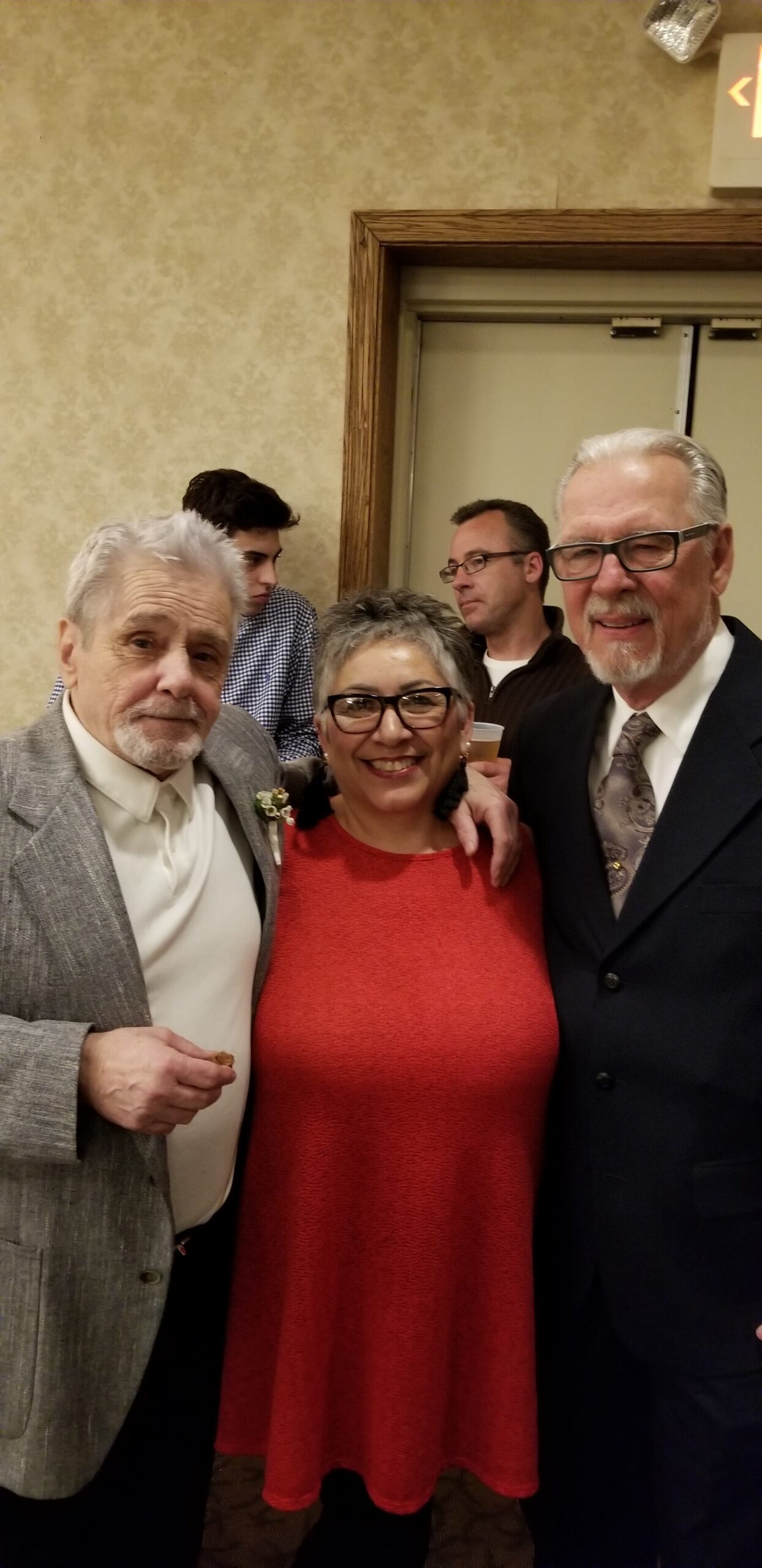 Three smiling adults posing for a photo at a formal event: an older man in a gray suit, a woman in a red dress, and a man in a dark suit with glasses.