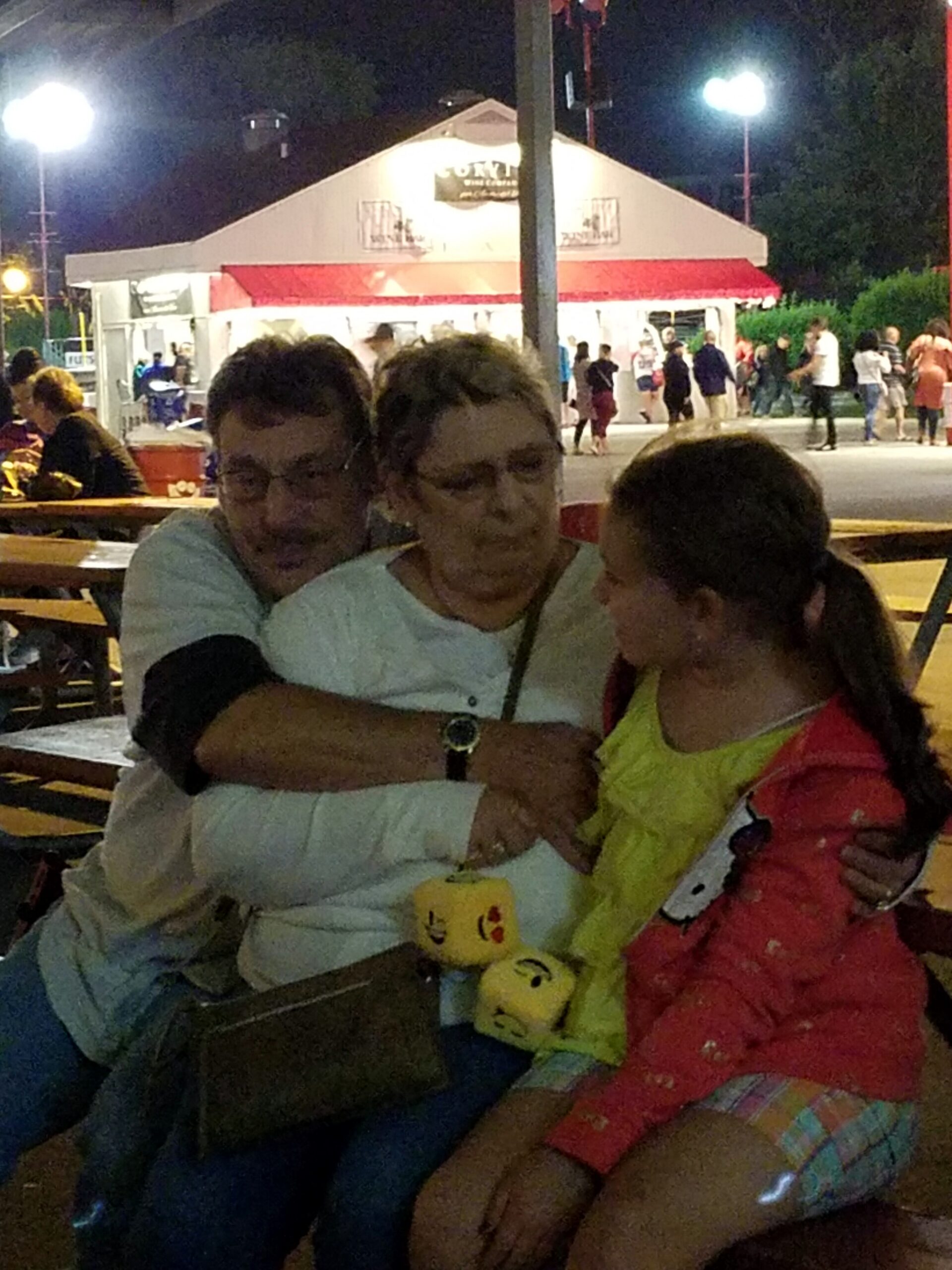 A couple sits at a picnic table at night, with a child in front, in an outdoor event space and a tent behind them.